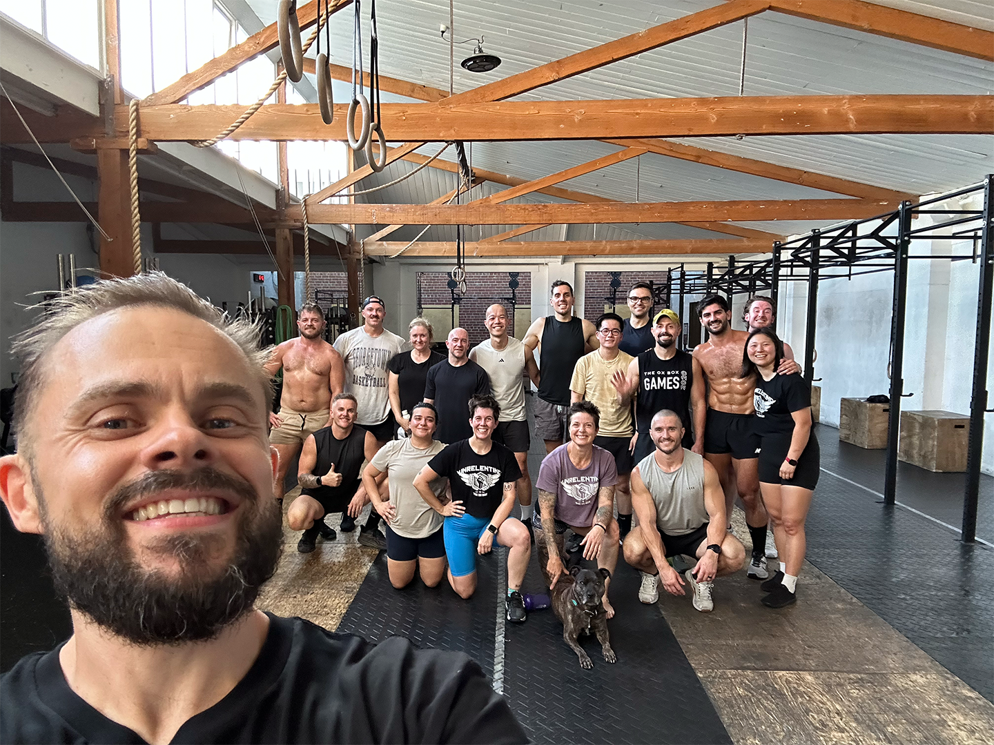 Group of people posing in a gym setting, wearing workout attire, with wooden beams and fitness equipment in the background.