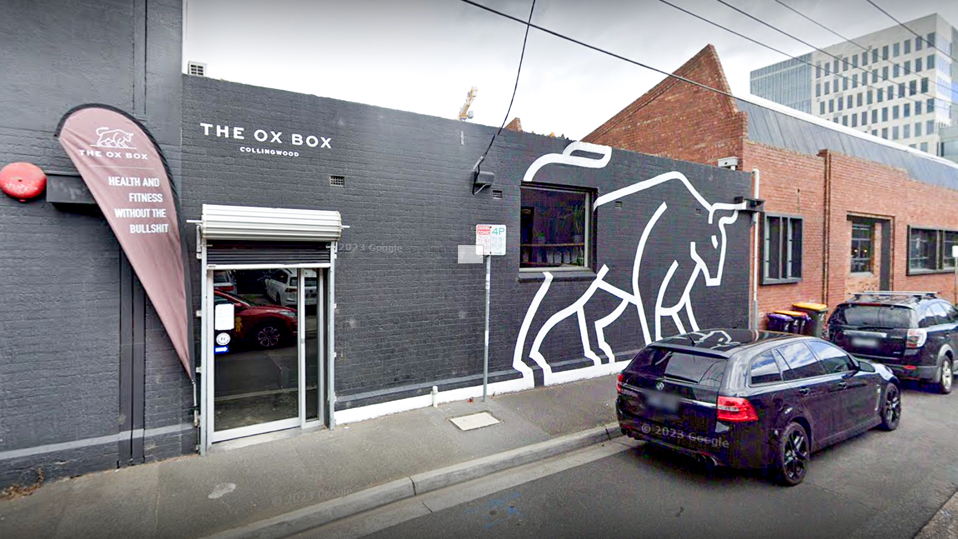Exterior of The Ox Box gym in Collingwood, featuring a large outline of an ox on the wall and a banner reading 'Health and fitness without the bullshit.' Several cars are parked outside the building.