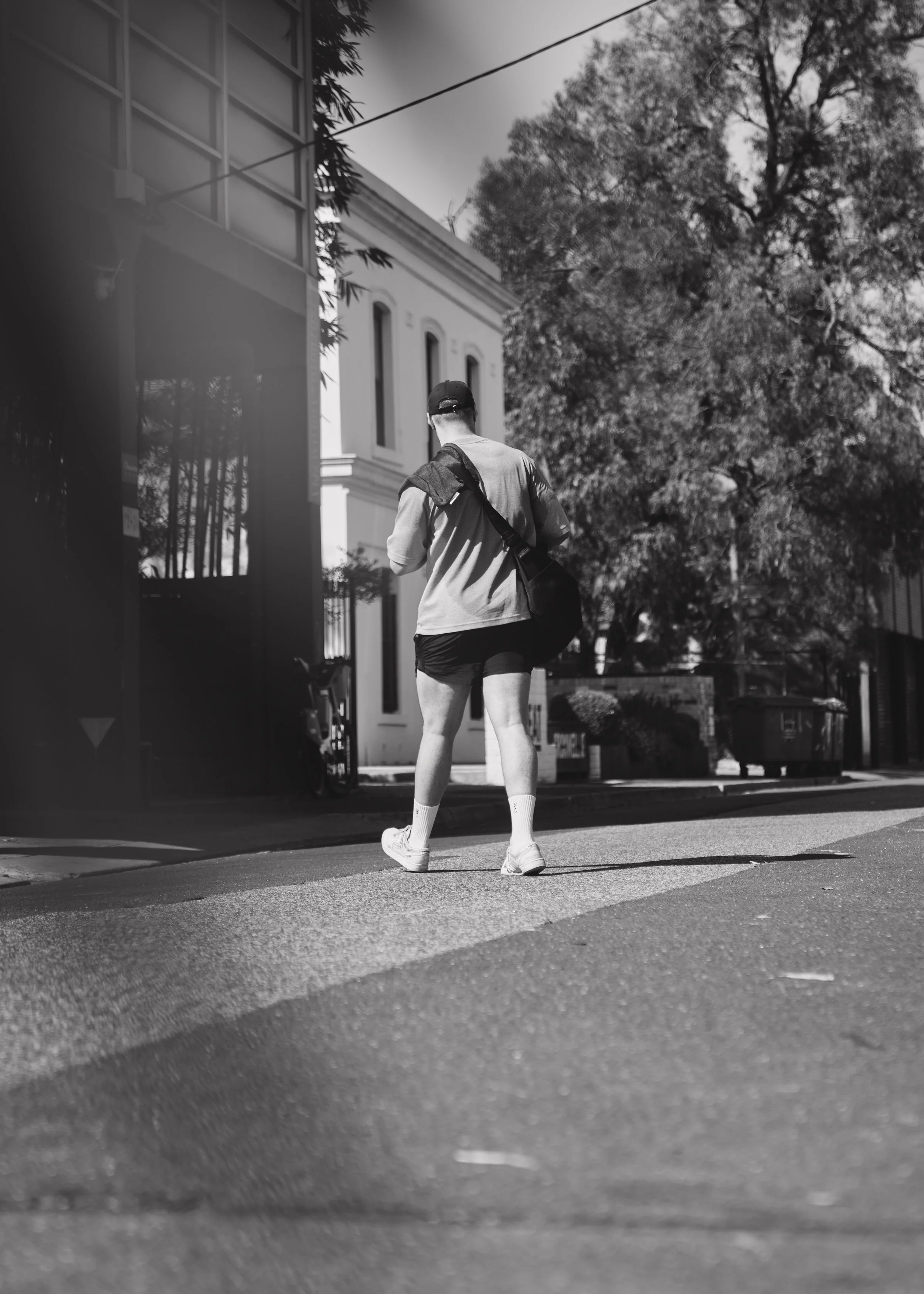 Black and white photo of a person walking on a sidewalk. They wear a cap, t-shirt, shorts, and sneakers, carrying a bag. Trees and buildings in the background.
