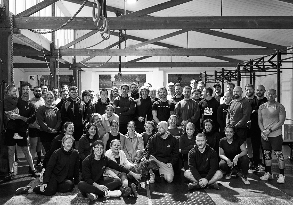 Group photo in a gym, featuring men and women of diverse ages, dressed in athletic attire, with gym equipment in the background. A small dog and a toddler are present.