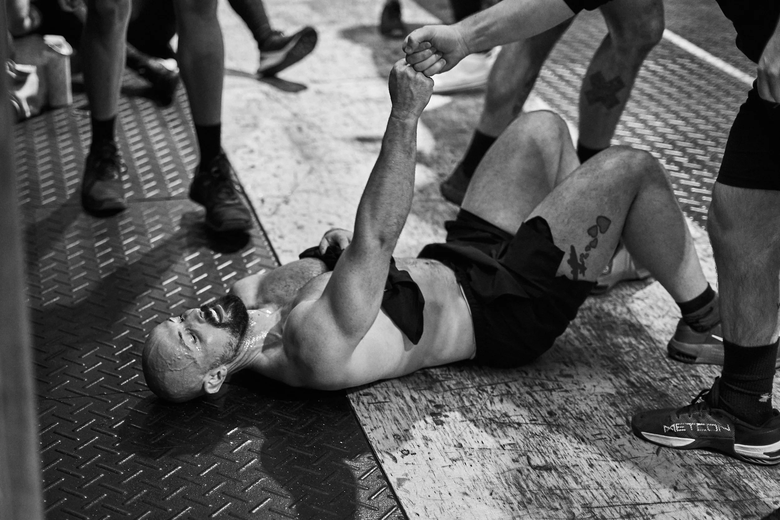A man lying on the gym floor, holding hands with another person, possibly after a workout. Other people and fitness equipment are visible in the background.