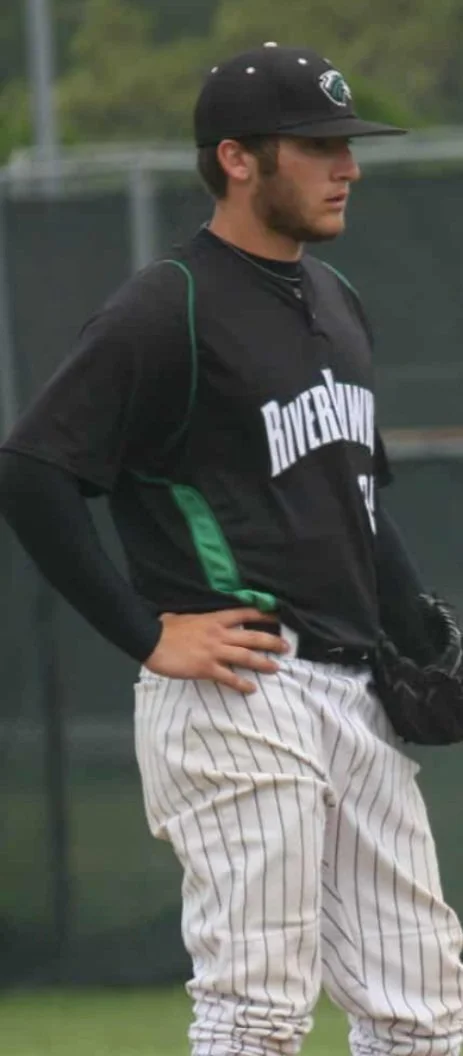 Young male baseball player in black and white striped uniform, wearing a black cap, standing with hand on hip, holding a baseball glove.