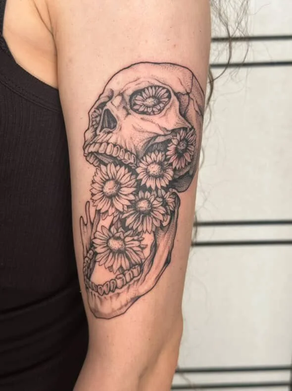 Tattoo of a human skull with flowers filling the inside of the skull on a person's upper arm.