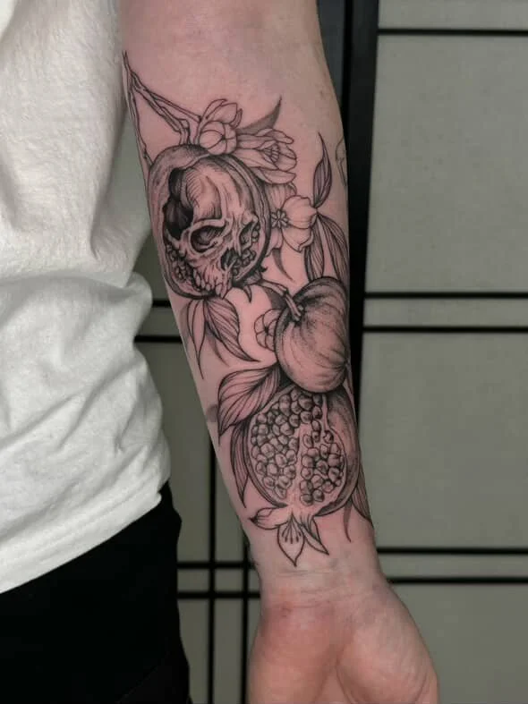 Black and gray tattoo of a skull, an apple, and a pomegranate with leaves on a person's forearm.