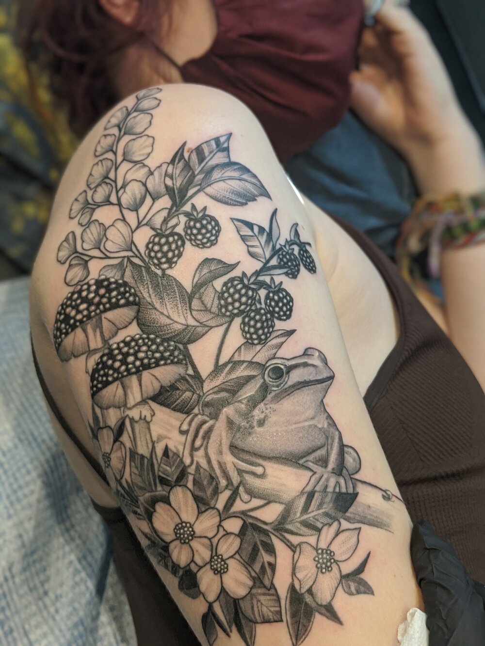 person with an illustrative nature tattoo with a frog, fern, mushrooms, and berries on their arm.