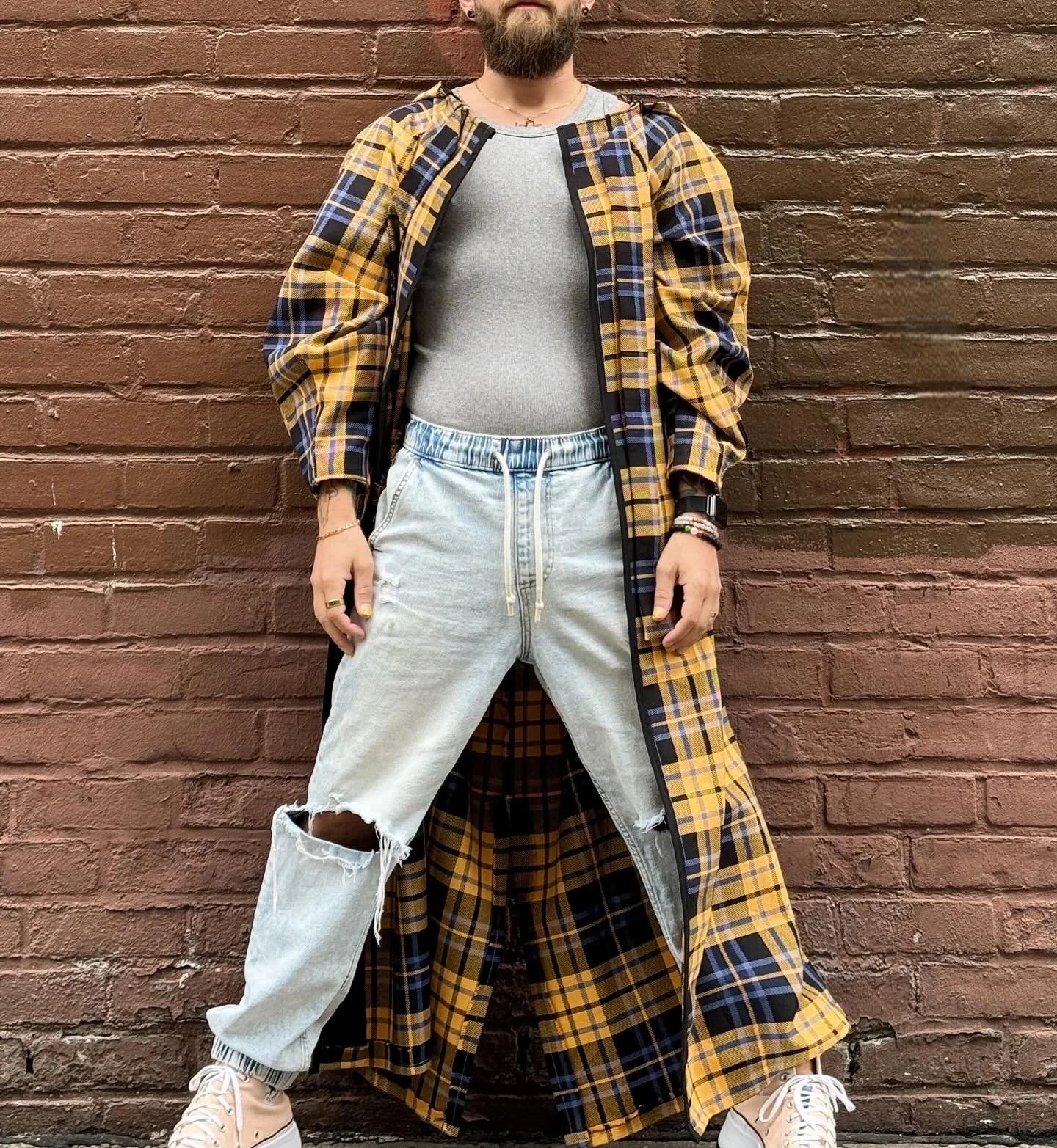 Person wearing a long yellow and black checkered coat, gray T-shirt, ripped light blue jeans, white sneakers, posing against a brick wall.