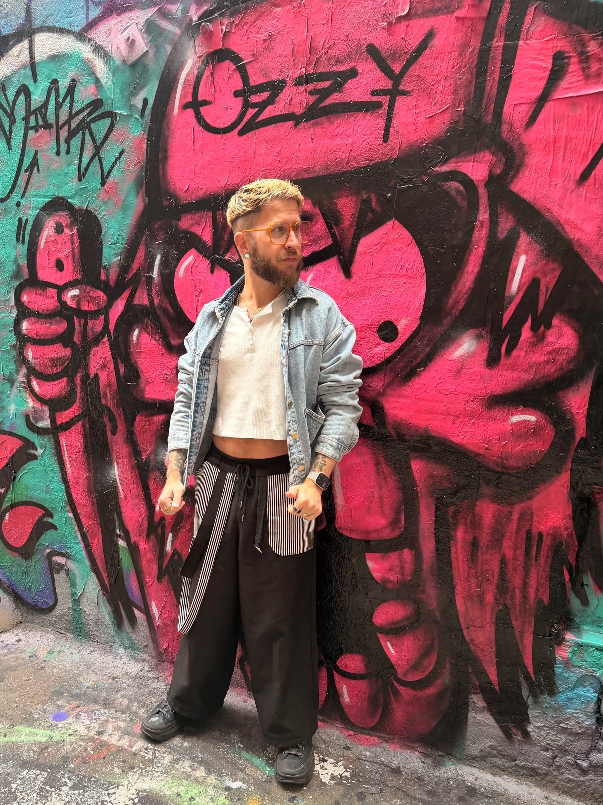 A person with short blond hair, glasses, earrings, and tattoos, wearing a light-colored crop top, denim jacket, and black pants, stands in front of a colorful graffiti mural on a brick wall. The graffiti features large pink and black abstract faces w