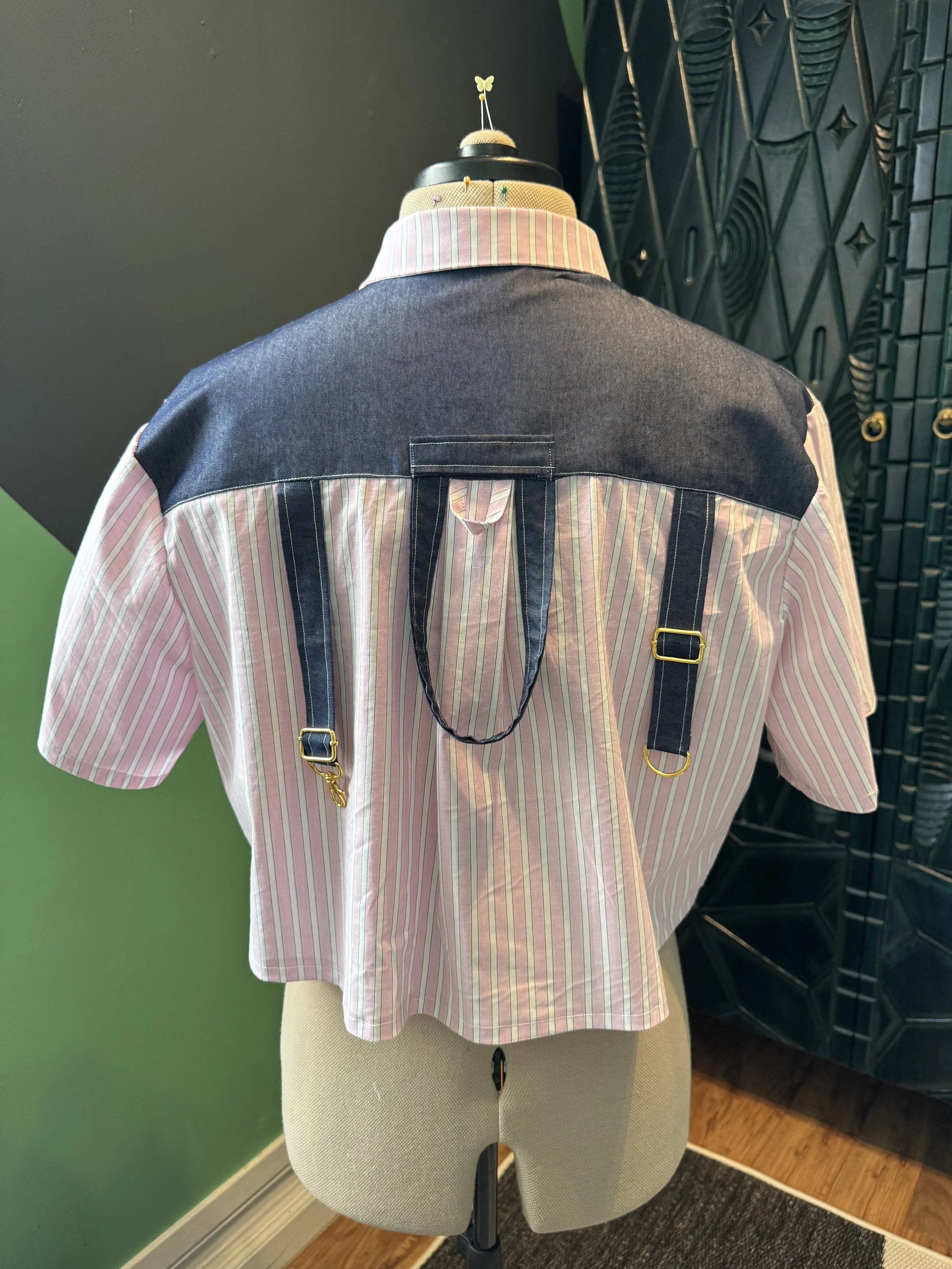 Back view of a mannequin wearing a shirt with red and white vertical stripes, black suspenders, and a black and beige vest. The background includes a green wall and a decorative black partition.