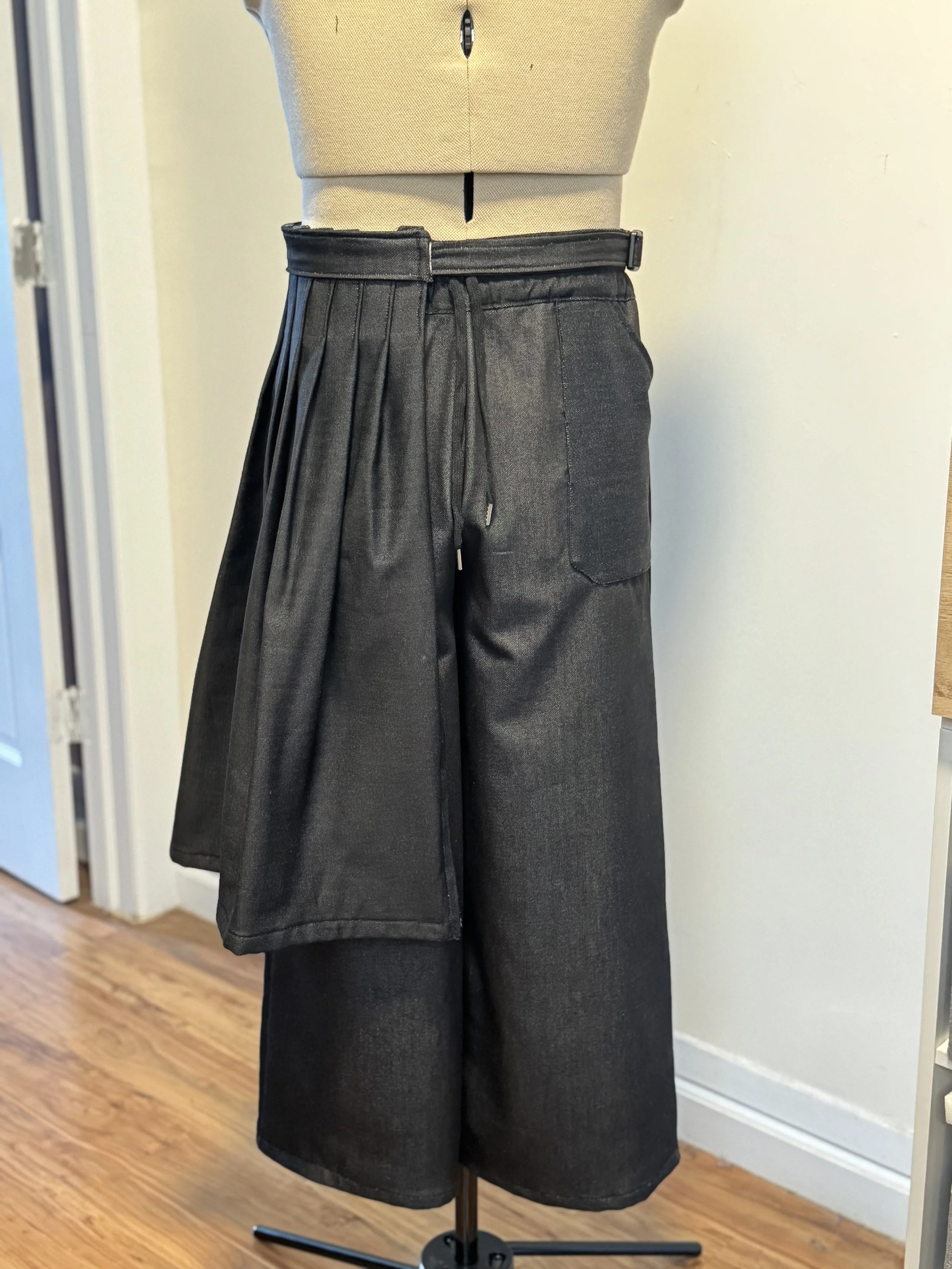 A mannequin wearing black pleated wide-leg pants with an elastic waistband, displayed on a black stand in a room with hardwood flooring and a light-colored wall.