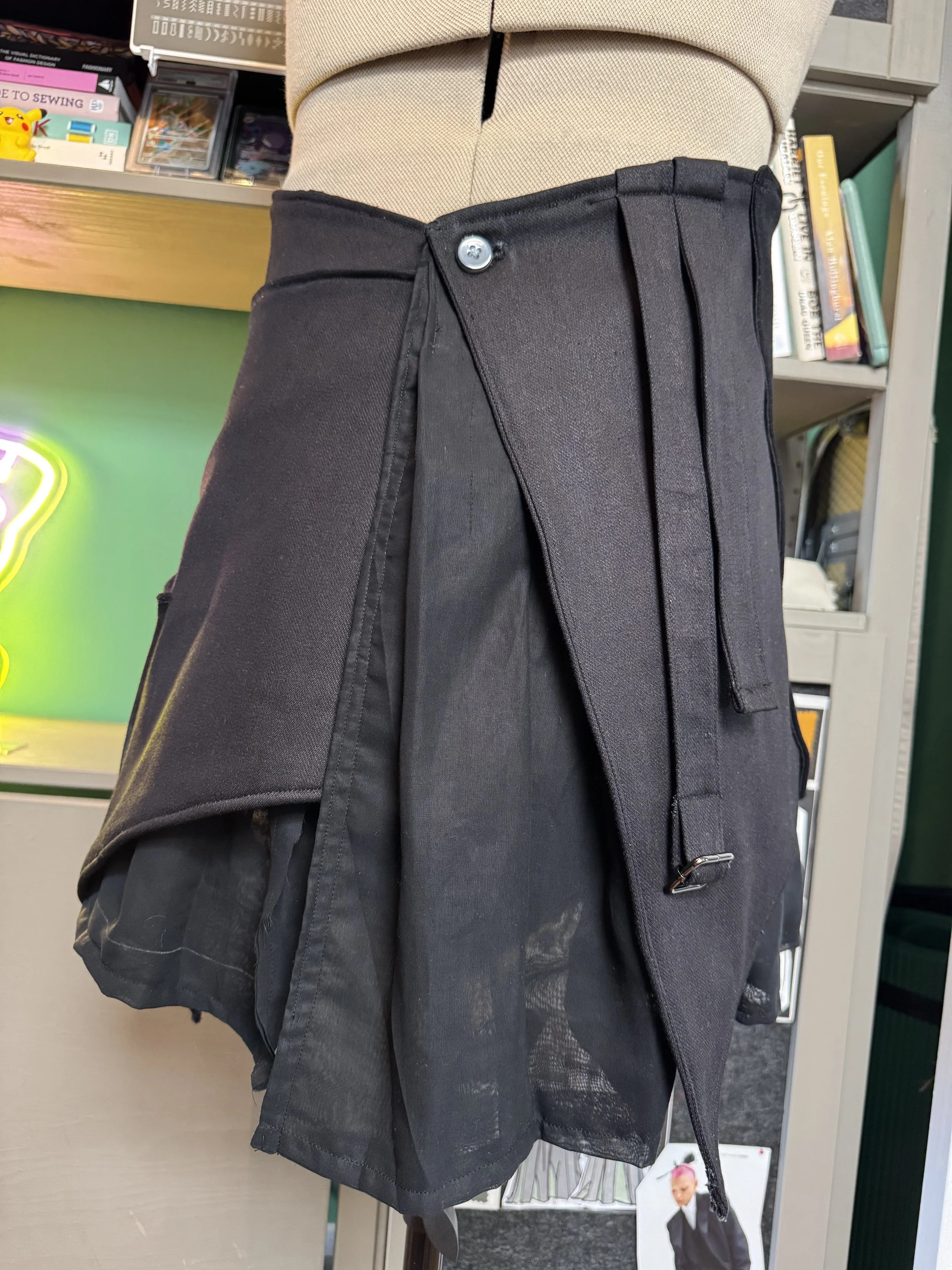 A mannequin wearing a black pleated skirt with a button and drawstring detail, displayed in a retail store with books and other items on shelves in the background.