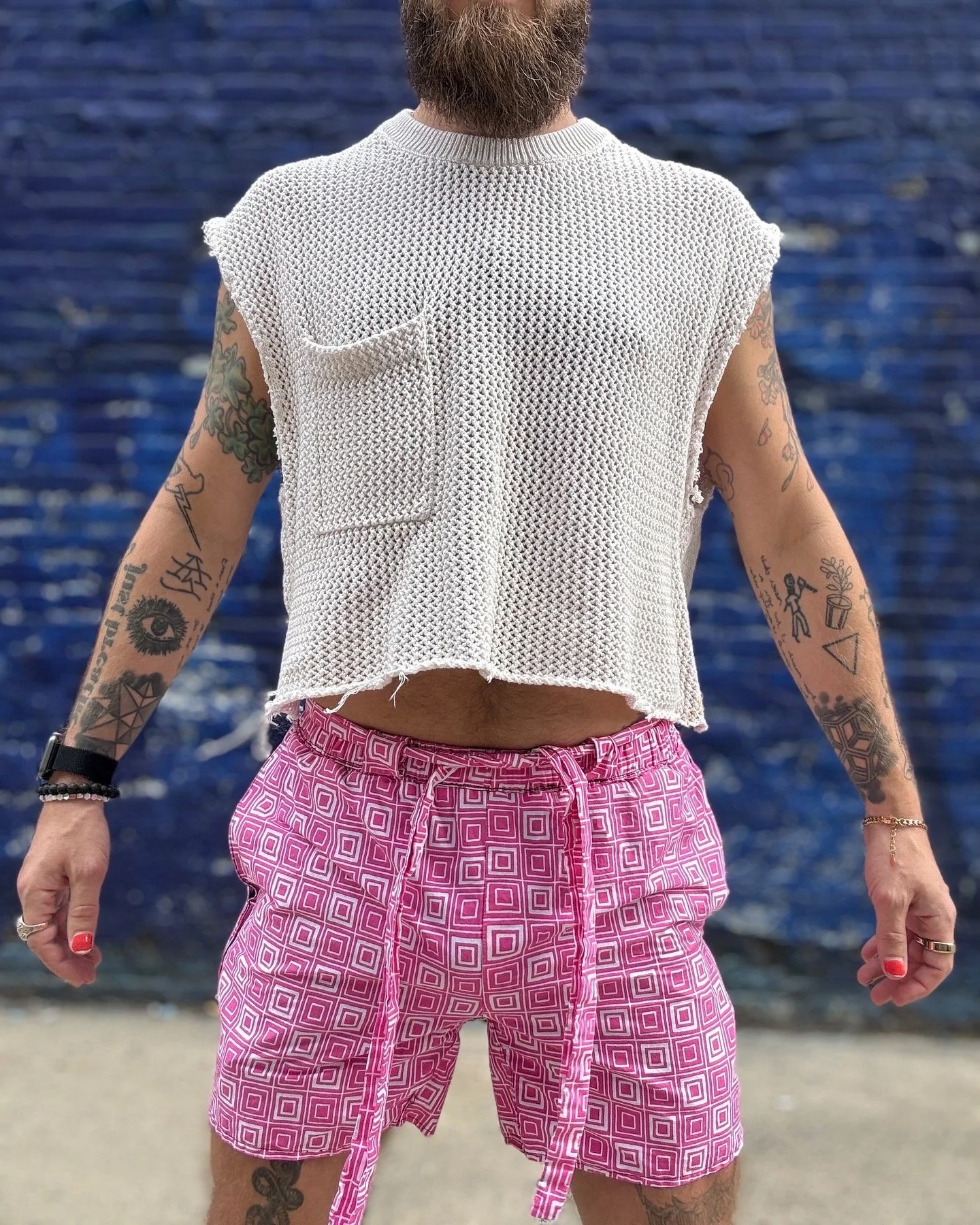 Person wearing a white mesh sleeveless top and pink geometric patterned shorts standing on a beach with ocean in the background.
