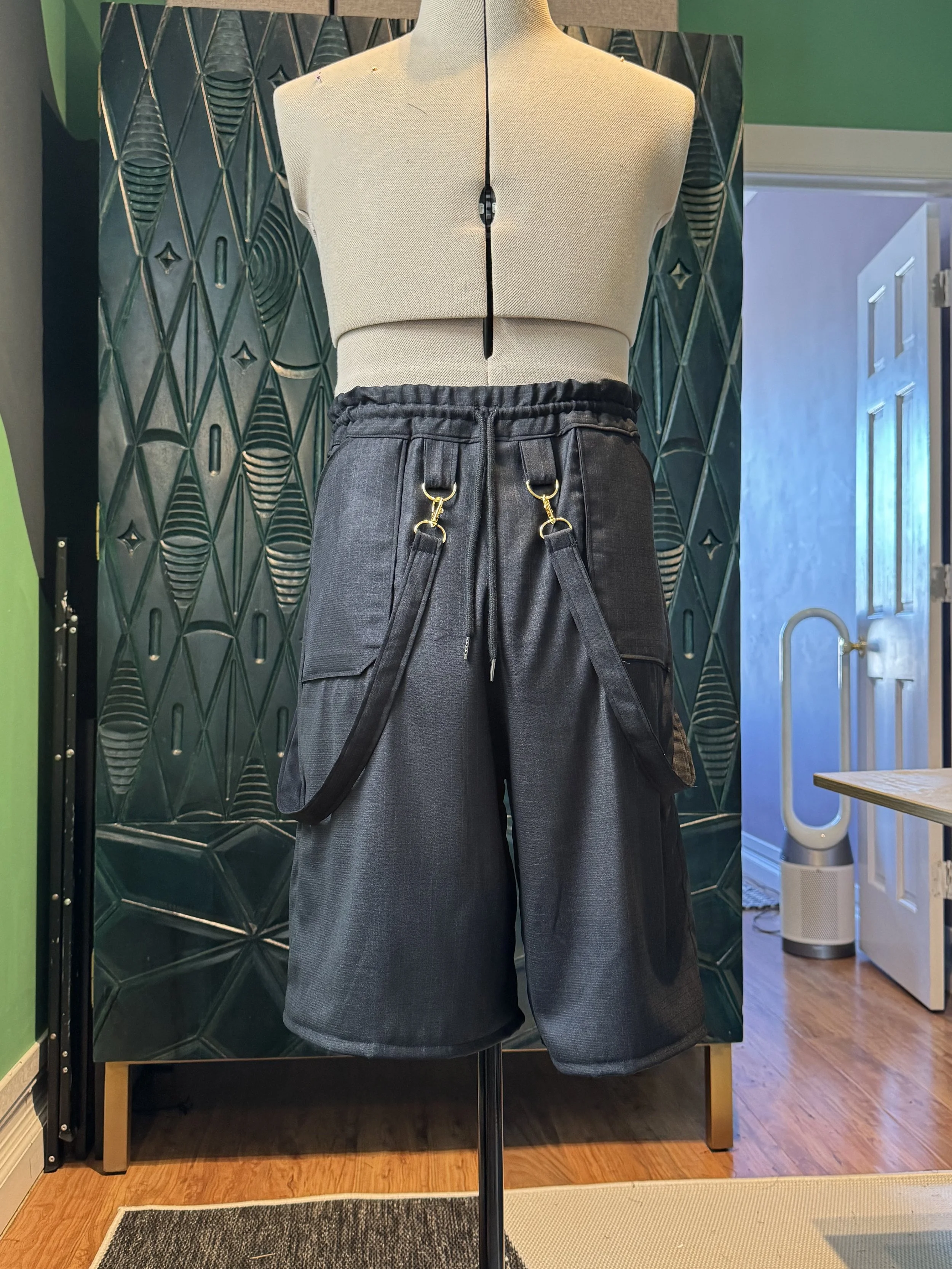 A mannequin torso on display with a beige sleeveless crop top and black pleated cargo pants with extra black straps hanging from the pants, in front of a decorative black tiled wall.