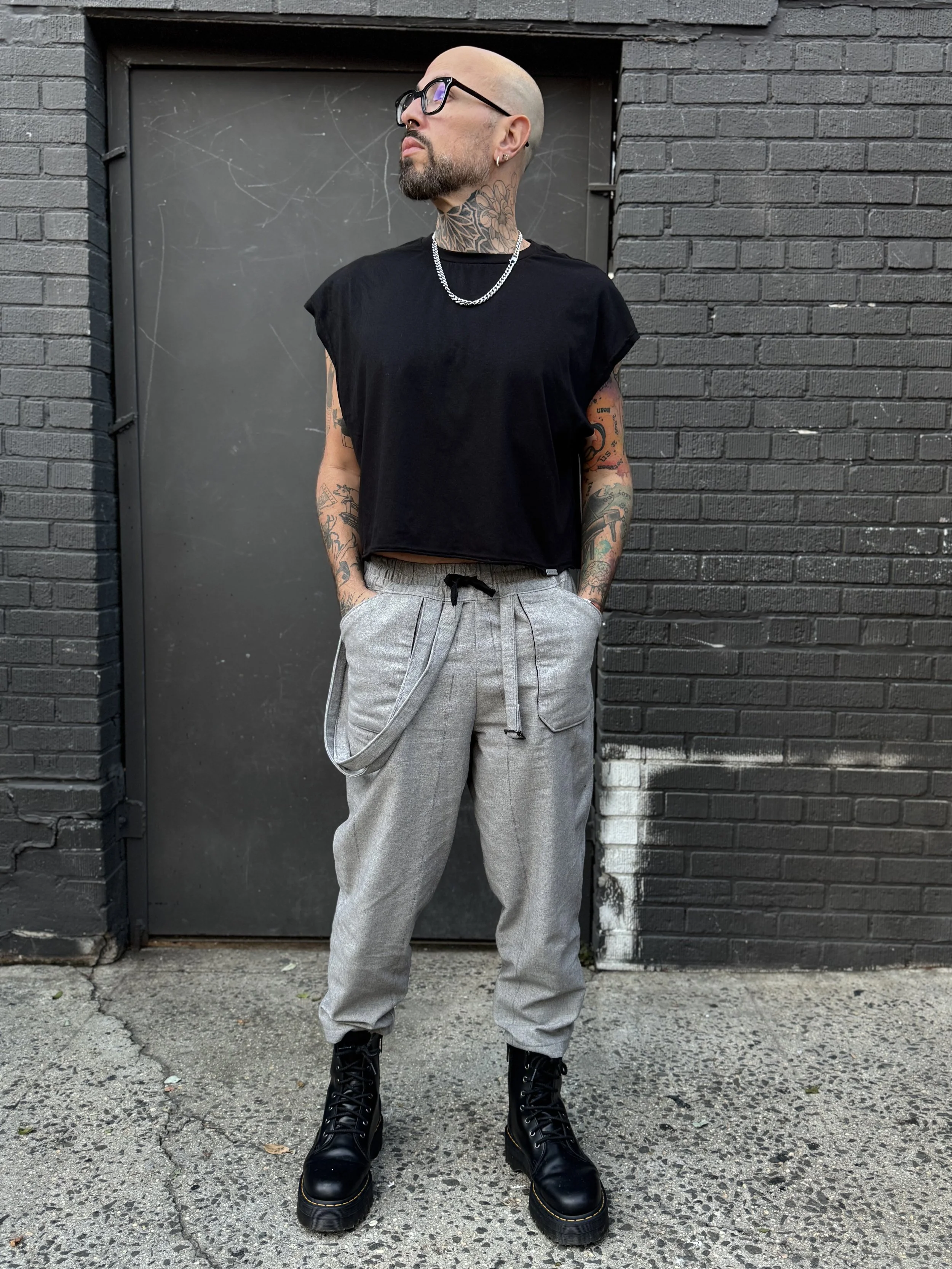 A man standing in front of a black brick wall and gray door, wearing black glasses, a black sleeveless top, gray baggy pants, black combat boots, silver jewelry, and tattoos on his arms and neck.