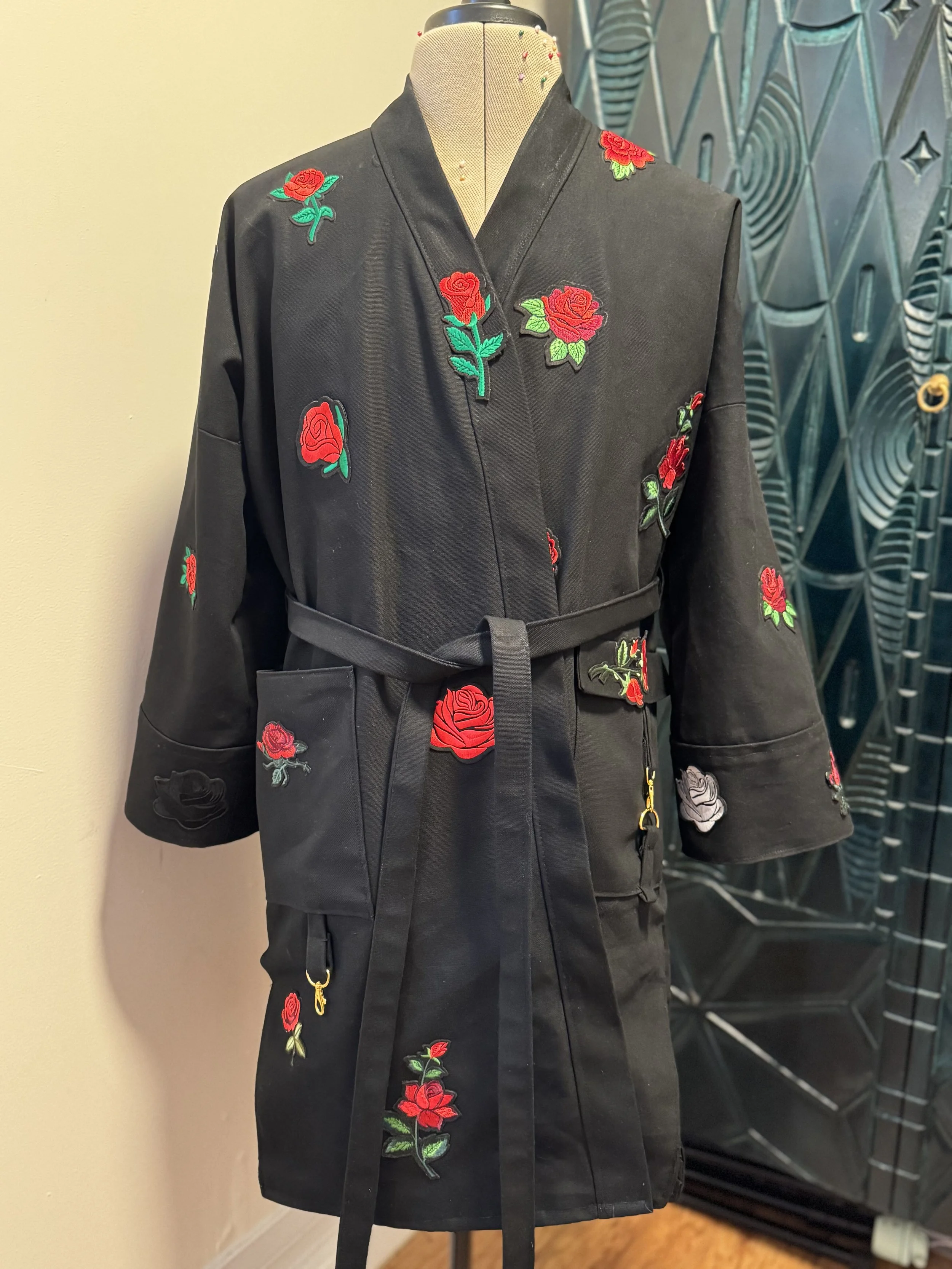 Black coat with embroidered red roses with green leaves on the sleeves, front, and side, displayed on a mannequin.