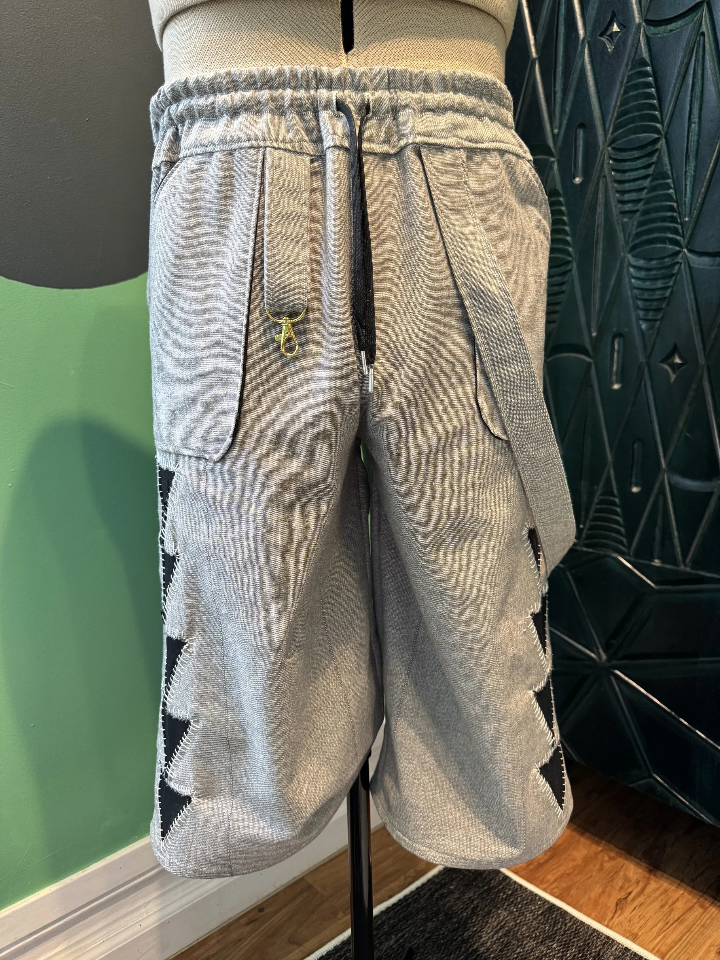 A mannequin dressed in gray shorts with zippers on the sides, standing in front of a black decorative wall panel and a green wall, with wooden flooring and a black rug underneath.