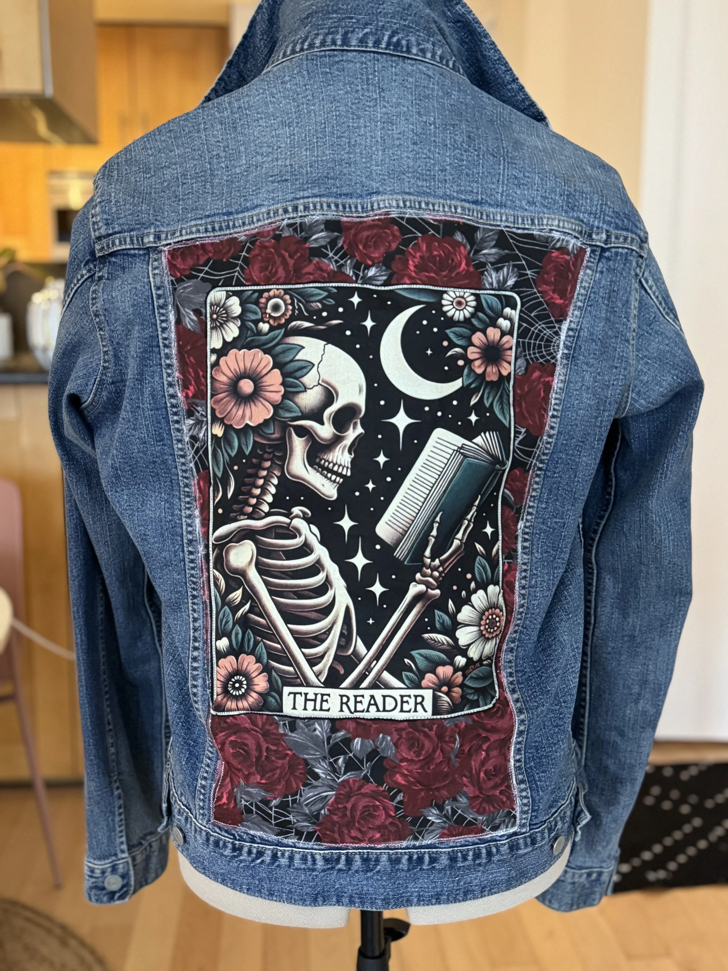 Denim jacket with a large patch on the back featuring a skeleton, flowers, a moon, and stars, with the text 'The Reader' at the bottom of the patch.