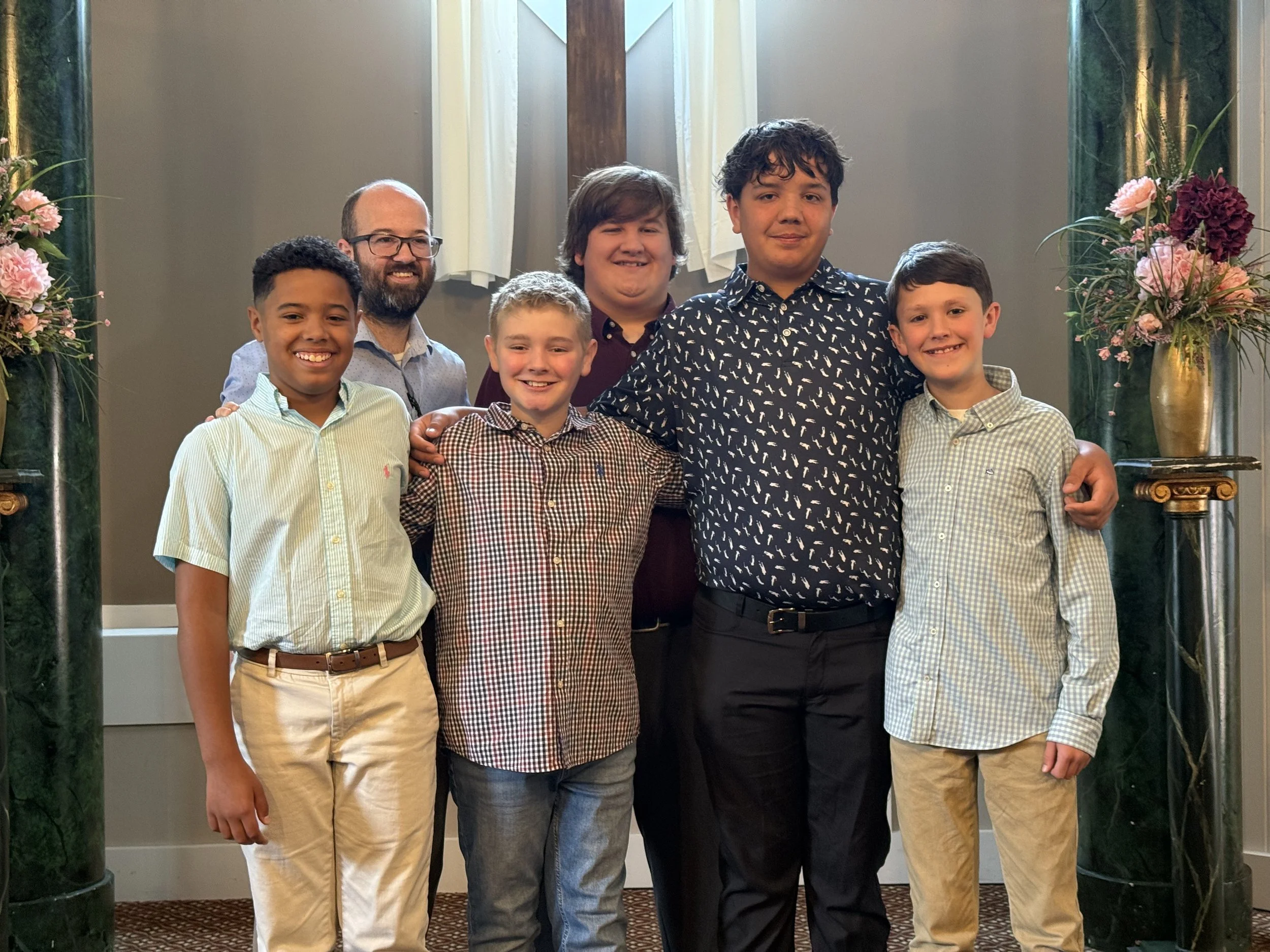 A group of seven boys and young men standing together, smiling, in front of a decorative backdrop with floral arrangements and columns.