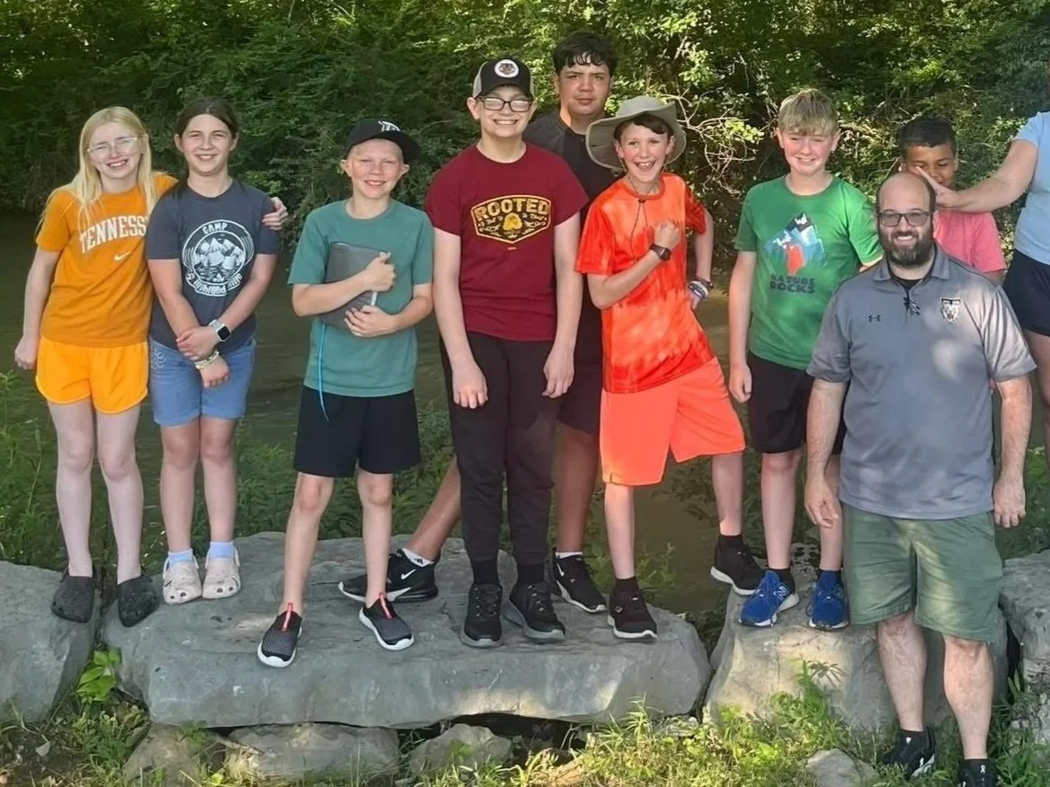 Group of children and two adults outdoors near a river, standing on rocks, smiling and posing for a photo in a wooded area.