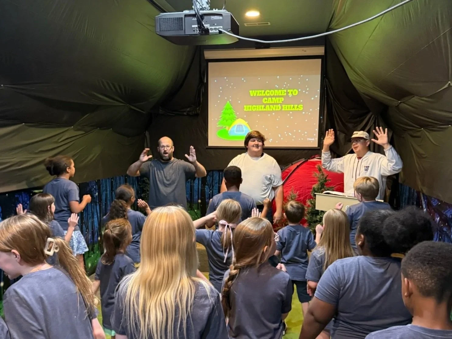 Children and adults gathered inside a tent or indoor shelter with a green black ceiling, participating in an activity or program. A screen at the front displays the message 'Welcome to Camp Highland Hills' with a graphic of a tent, trees, and stars. 