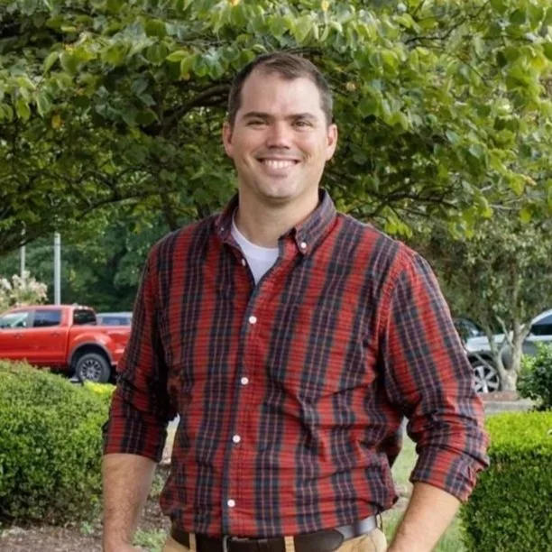A man in a red and black plaid shirt smiling outdoors in front of green trees and bushes, with parked cars in the background.