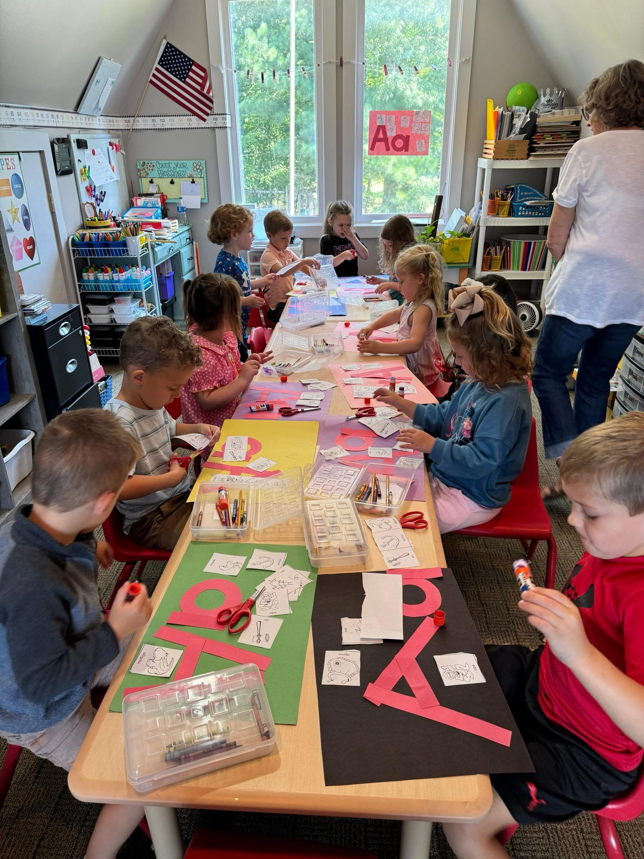 Classroom with children engaged in craft activities at a long table with paper, scissors, glue, and markers. A teacher stands nearby. The classroom is decorated with an American flag, colorful posters, and supplies on shelves.