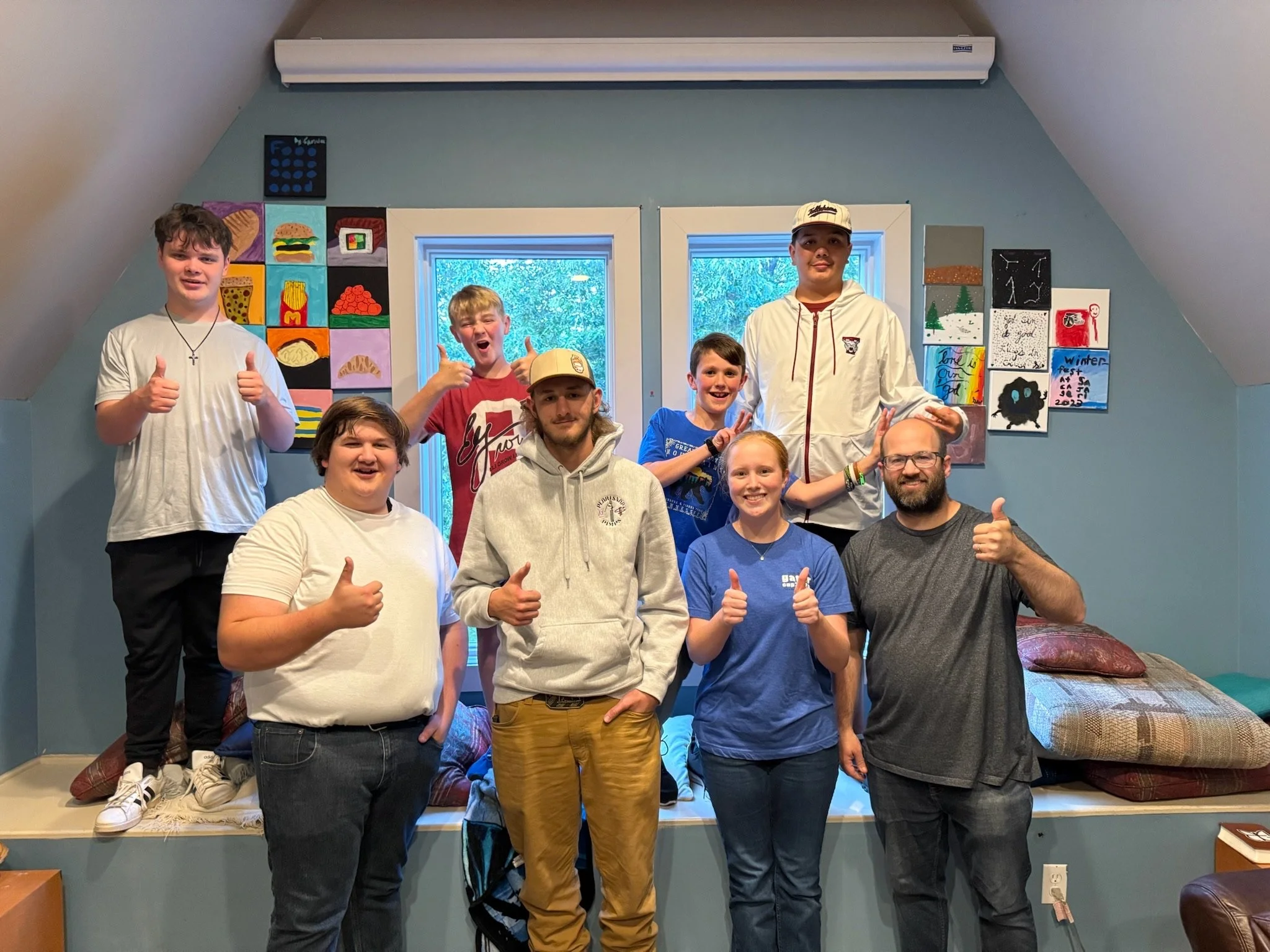 Group of nine people, mostly children and young adults, standing in a room with blue walls, giving thumbs-up gestures and smiling. Behind them are two windows and colorful artwork on the walls.