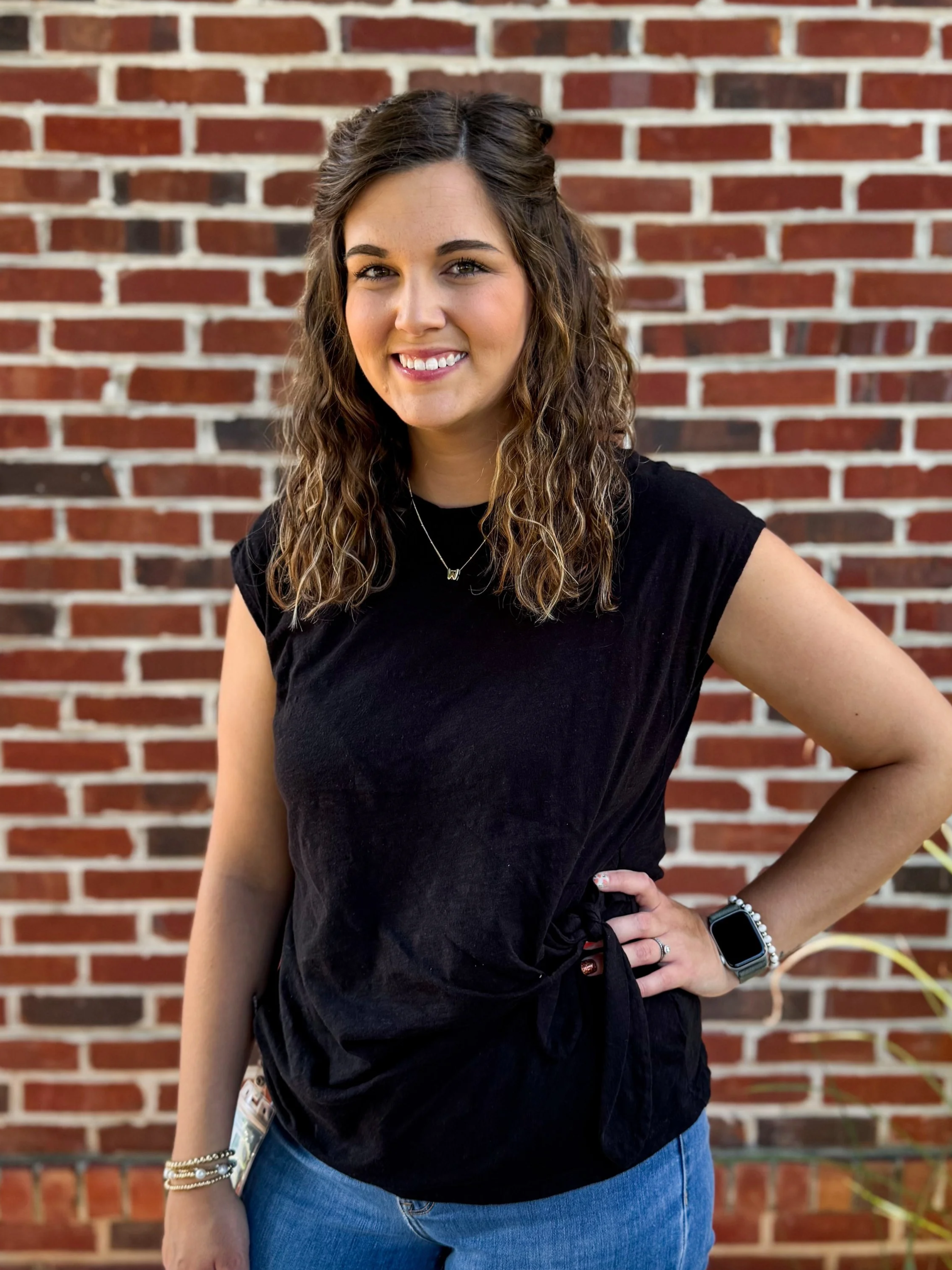 A young woman with wavy brown hair, wearing a black sleeveless top, blue jeans, and a smartwatch, standing in front of a red brick wall and smiling at the camera.