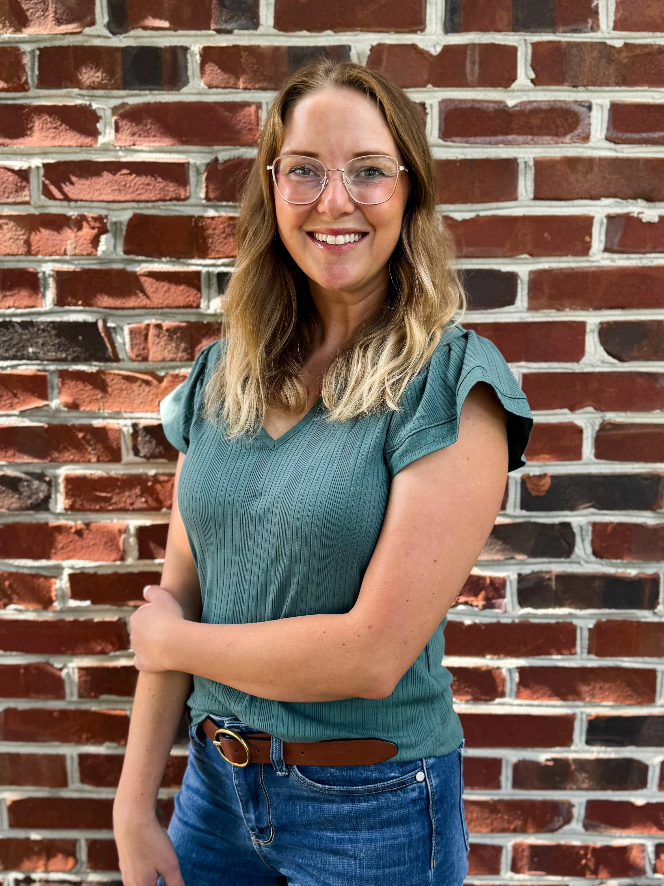 A woman with blonde, wavy hair, glasses, and a green ribbed sleeveless top standing in front of a red brick wall, smiling at the camera.