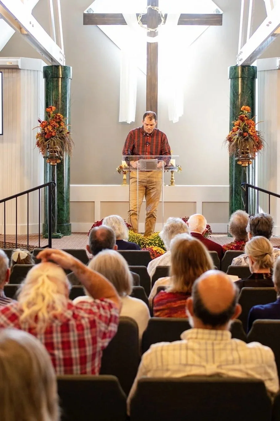 A man stands at a transparent lectern in front of a cross, speaking to an audience of elderly people in a church. The setting is decorated with floral arrangements and there is a bright light shining through white curtains behind him.