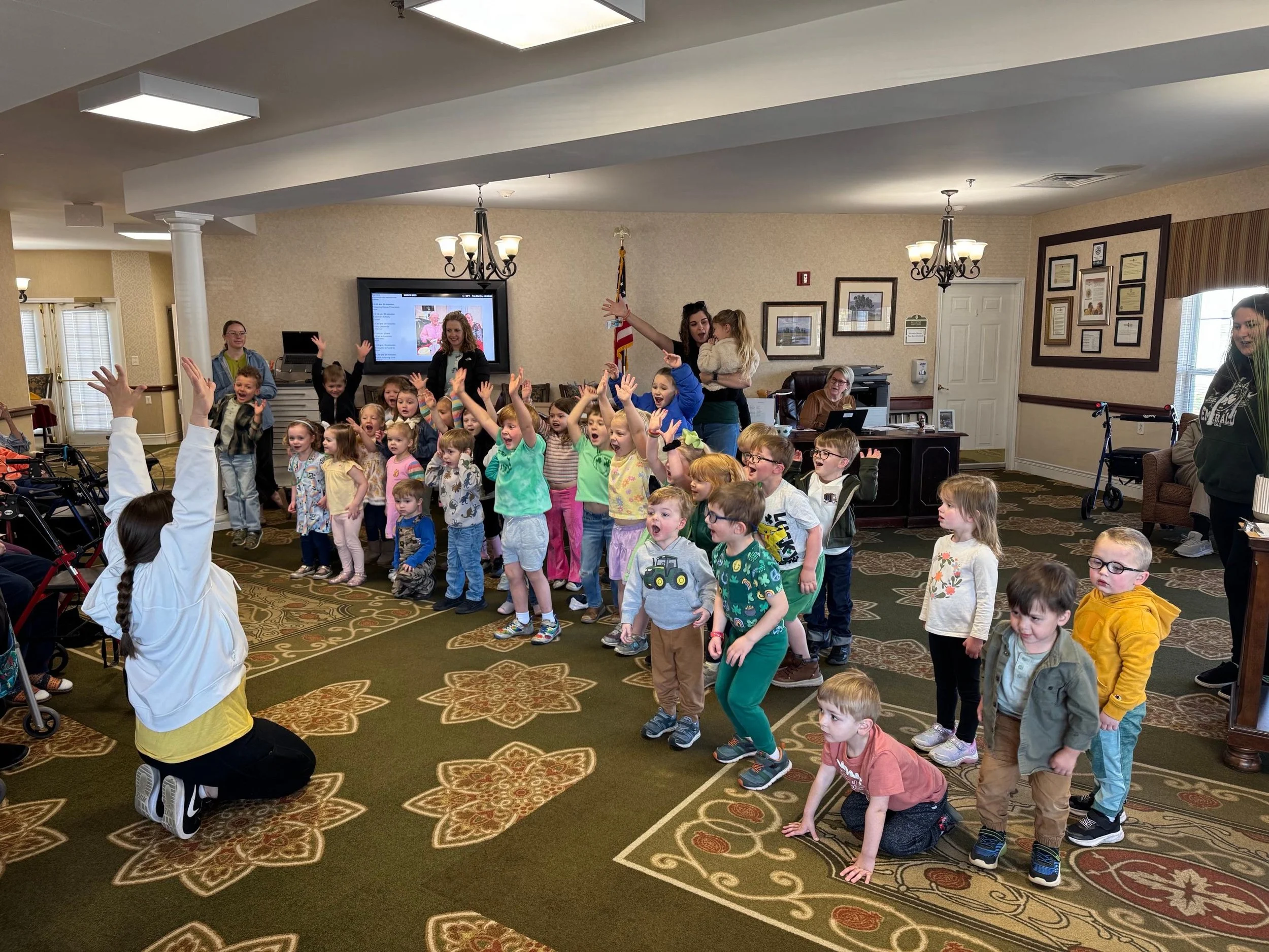 A group of children participating in an activity led by a woman in a large room, with adults observing. Some children have their hands raised, and a woman is holding a child.