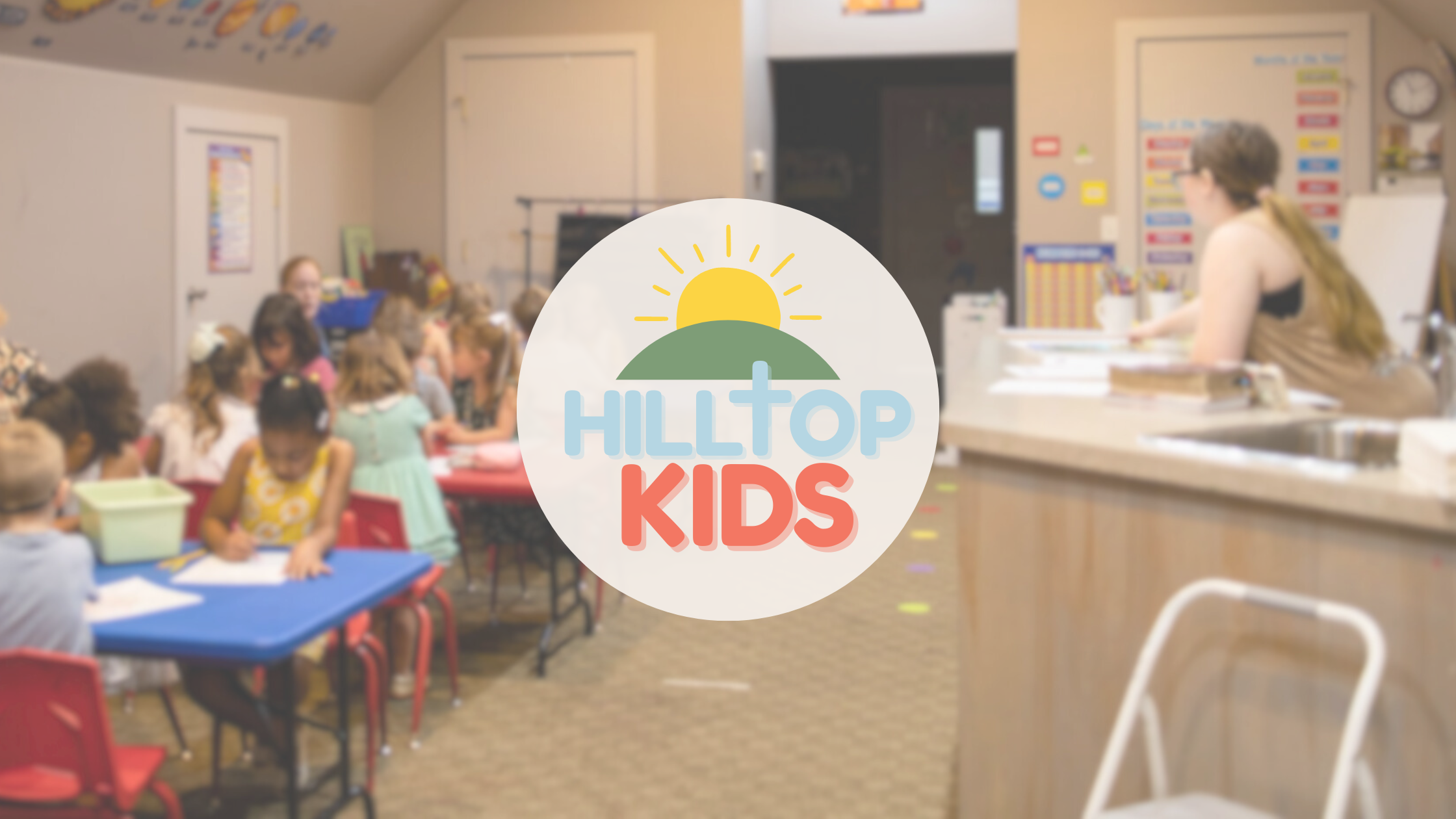 Classroom filled with young children sitting at tables and working on activities, with a teacher at the front, and a colorful logo in the center reading 'HILLTOP KIDS' with a sun and hill graphic.