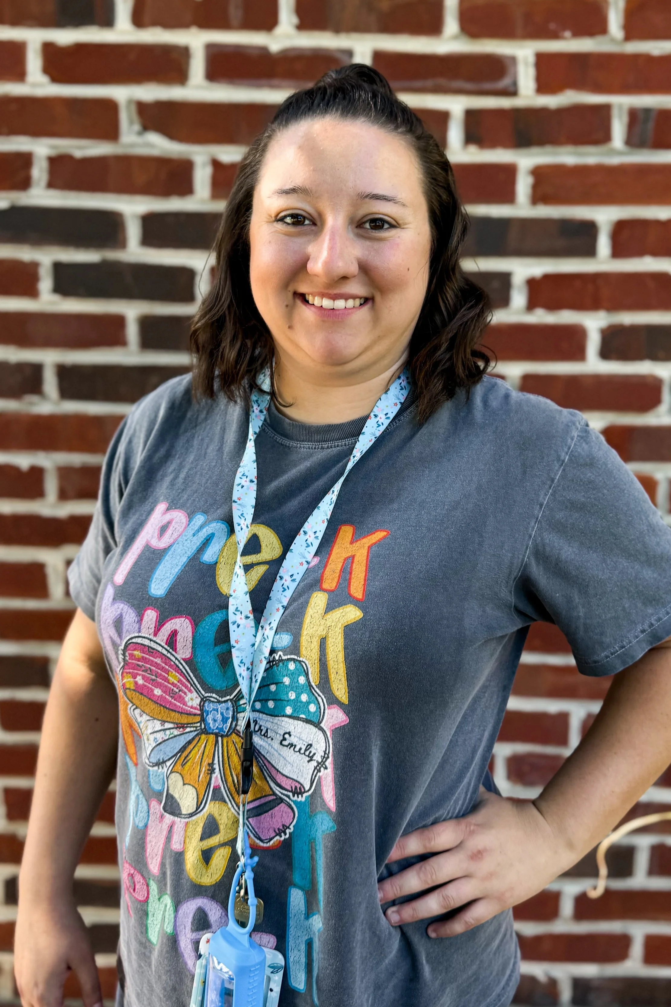 A woman with dark brown, shoulder-length, wavy hair smiling and standing in front of a brick wall. She is wearing a gray t-shirt with colorful text and a butterfly graphic, a lanyard with a small blue item attached, and has her right hand on her hip.