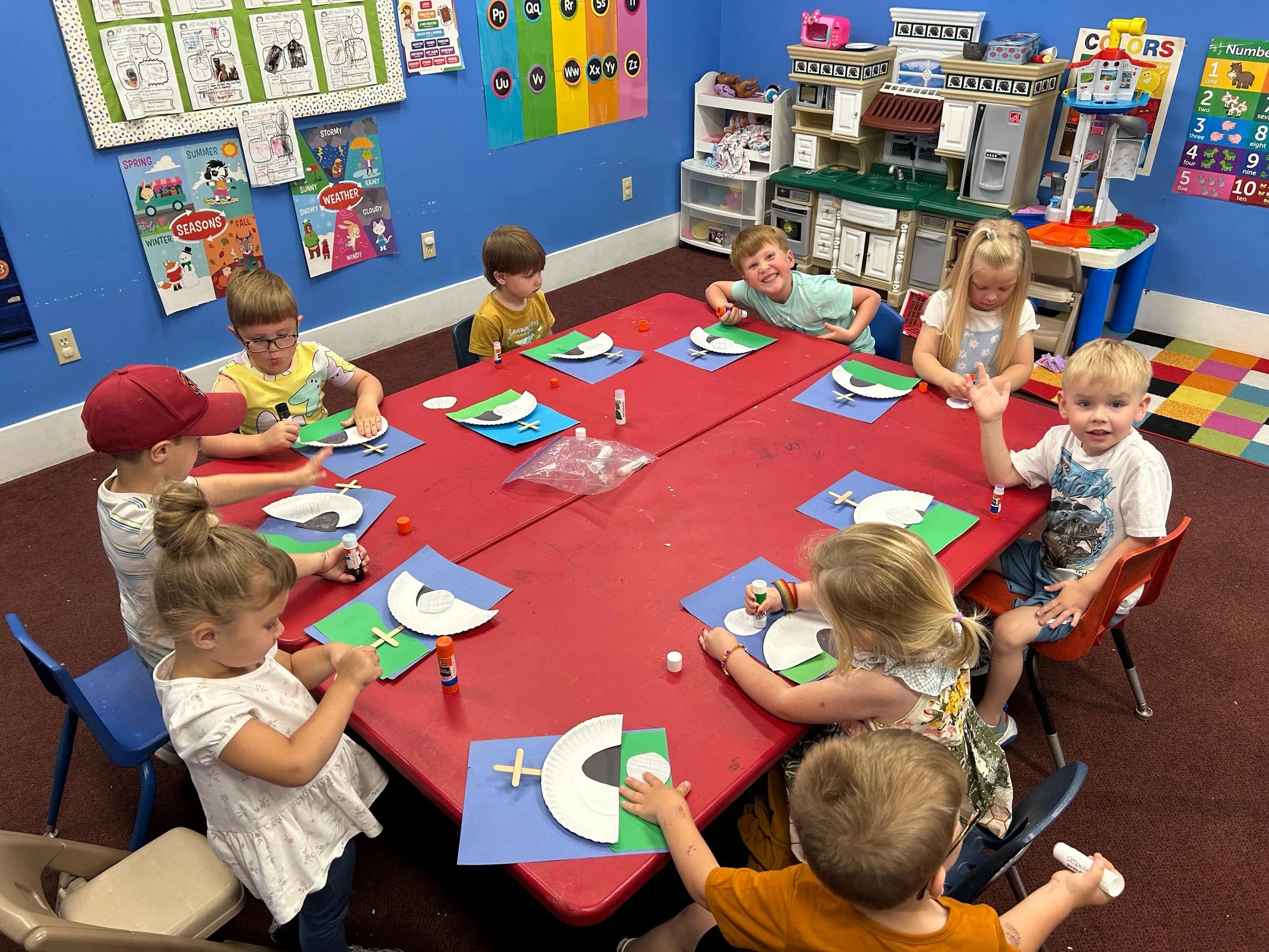 Children seated around a red table in a classroom, creating craft projects with paper, glue, and sticks, with colorful educational posters on the blue walls.