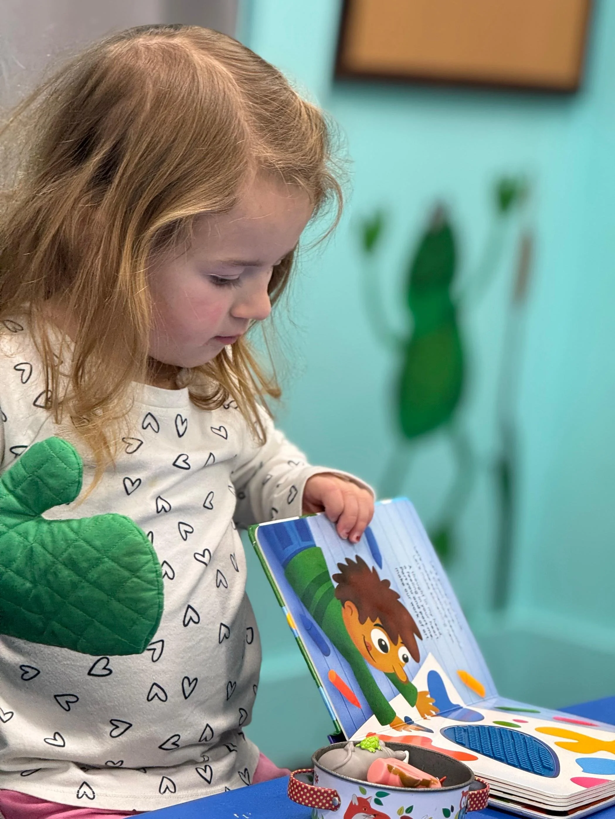 A young girl with red hair, wearing a white shirt with black hearts and green quilted mittens, looks at an open children's book with colorful illustrations. A decorative tin container with toys or figurines is on the table in front of her, and the ba