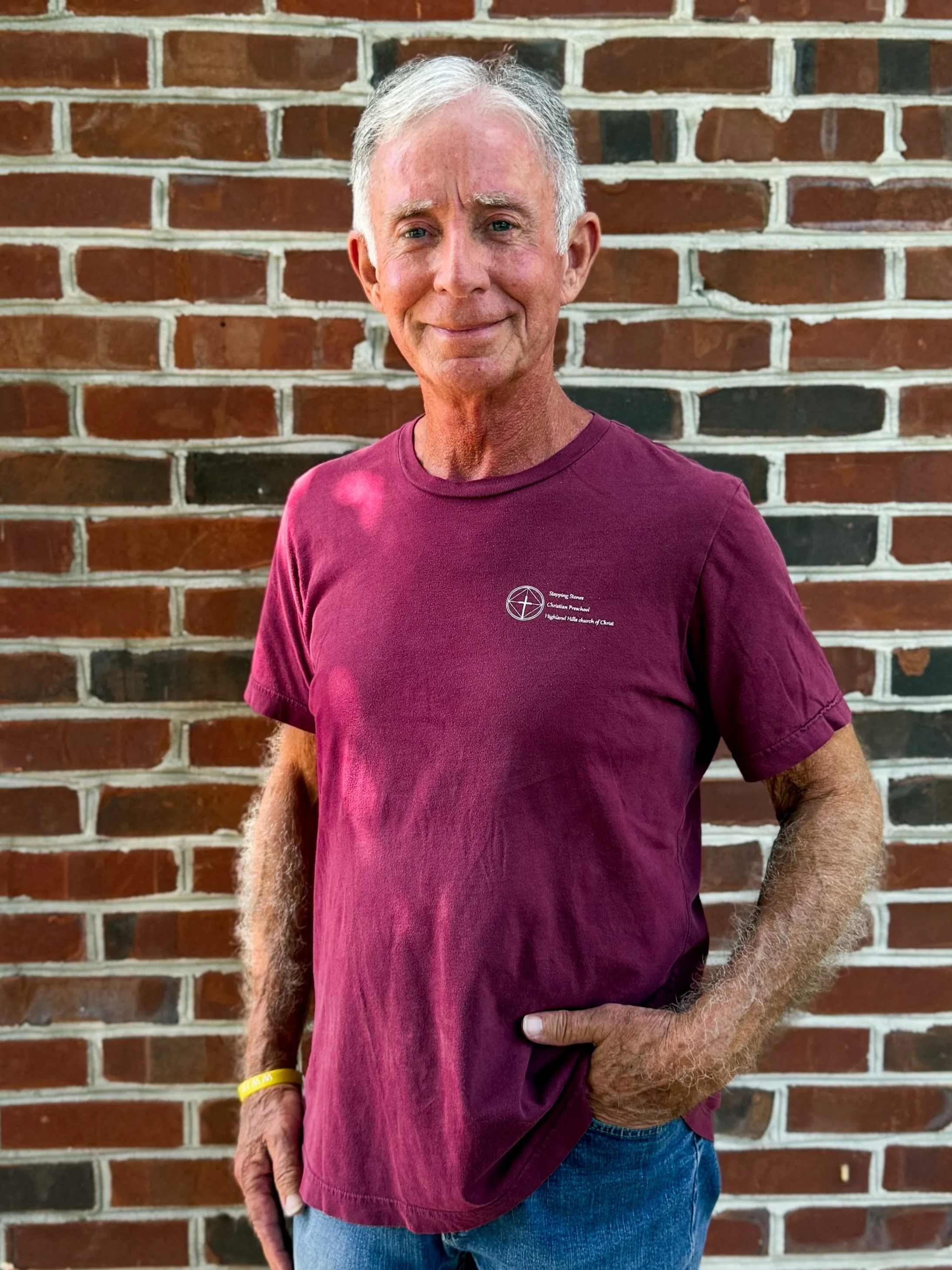 A smiling elderly man with gray hair, wearing a maroon t-shirt and blue jeans, standing outdoors in front of a brick wall.