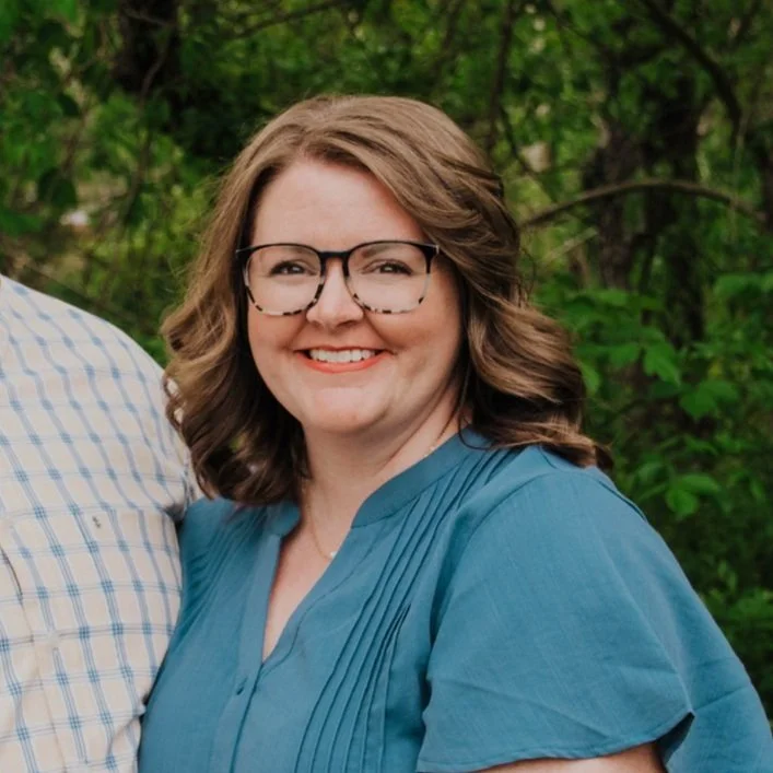 A woman with shoulder-length brown hair, wearing glasses and a blue blouse, smiling outdoors with greenery in the background.