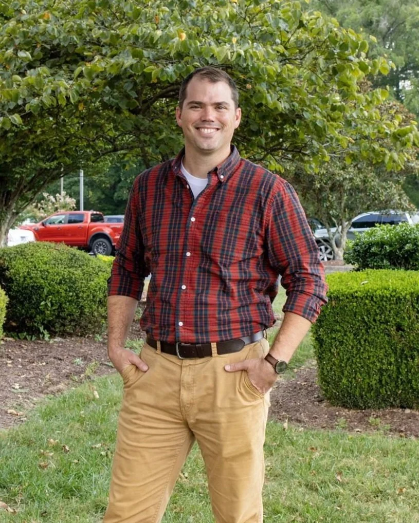 A smiling man wearing a red and black plaid shirt and tan pants standing outdoors in front of green shrubs and trees, with cars parked in the background.