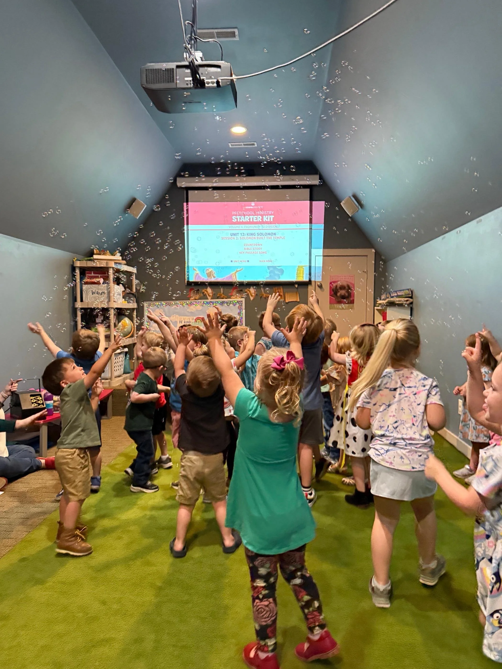 Children dancing and playing in a classroom with a screen displaying educational content at the front.