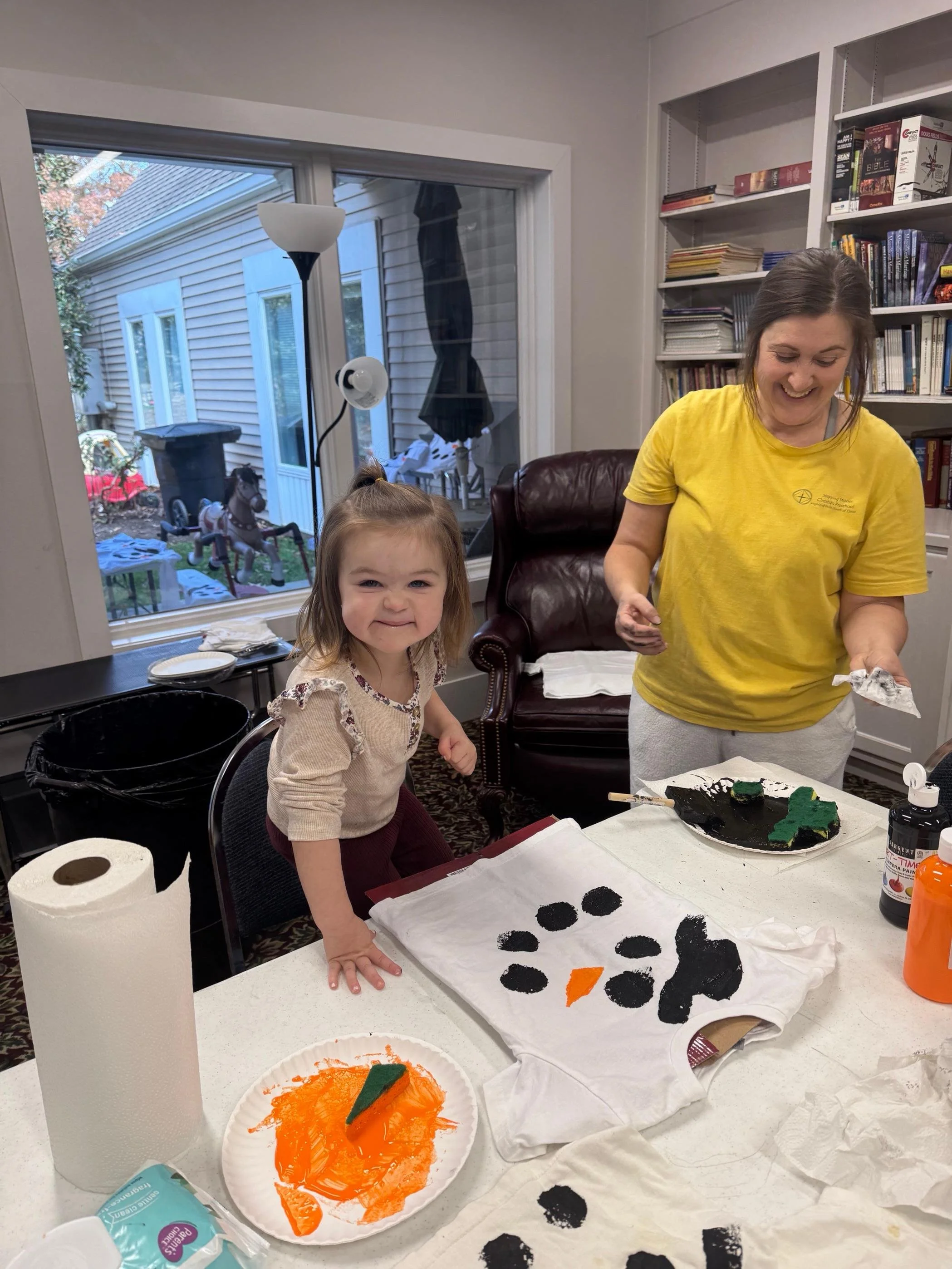 A woman and a young girl smiling and working on a craft project at a table, creating a snowman with black and orange paper. The table has painting supplies and paper towels, with a window showing outdoor toys and house siding outside.
