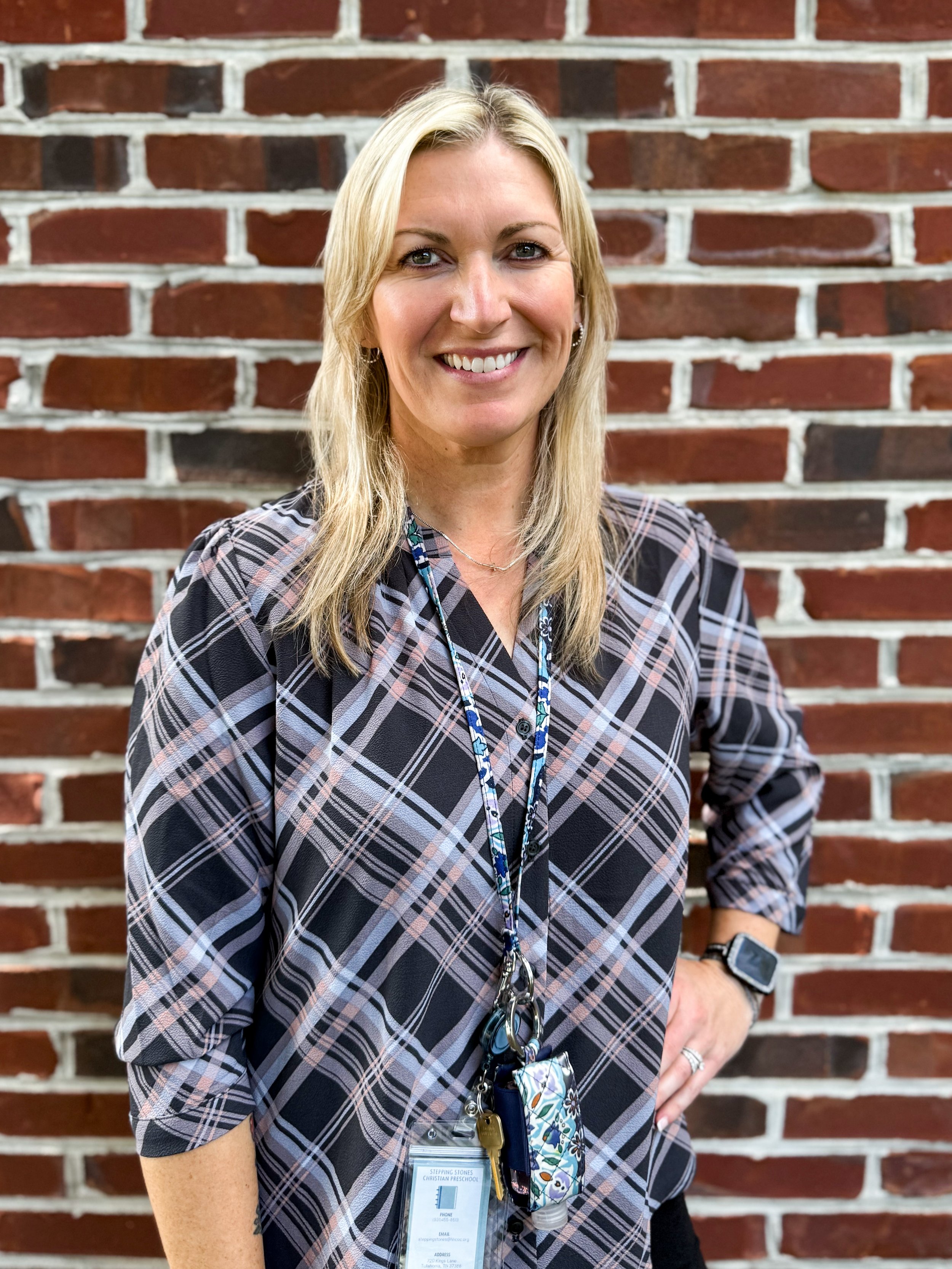 A smiling woman with blonde hair standing in front of a brick wall, wearing a plaid shirt, a lanyard with keys and a badge, and a smartwatch.