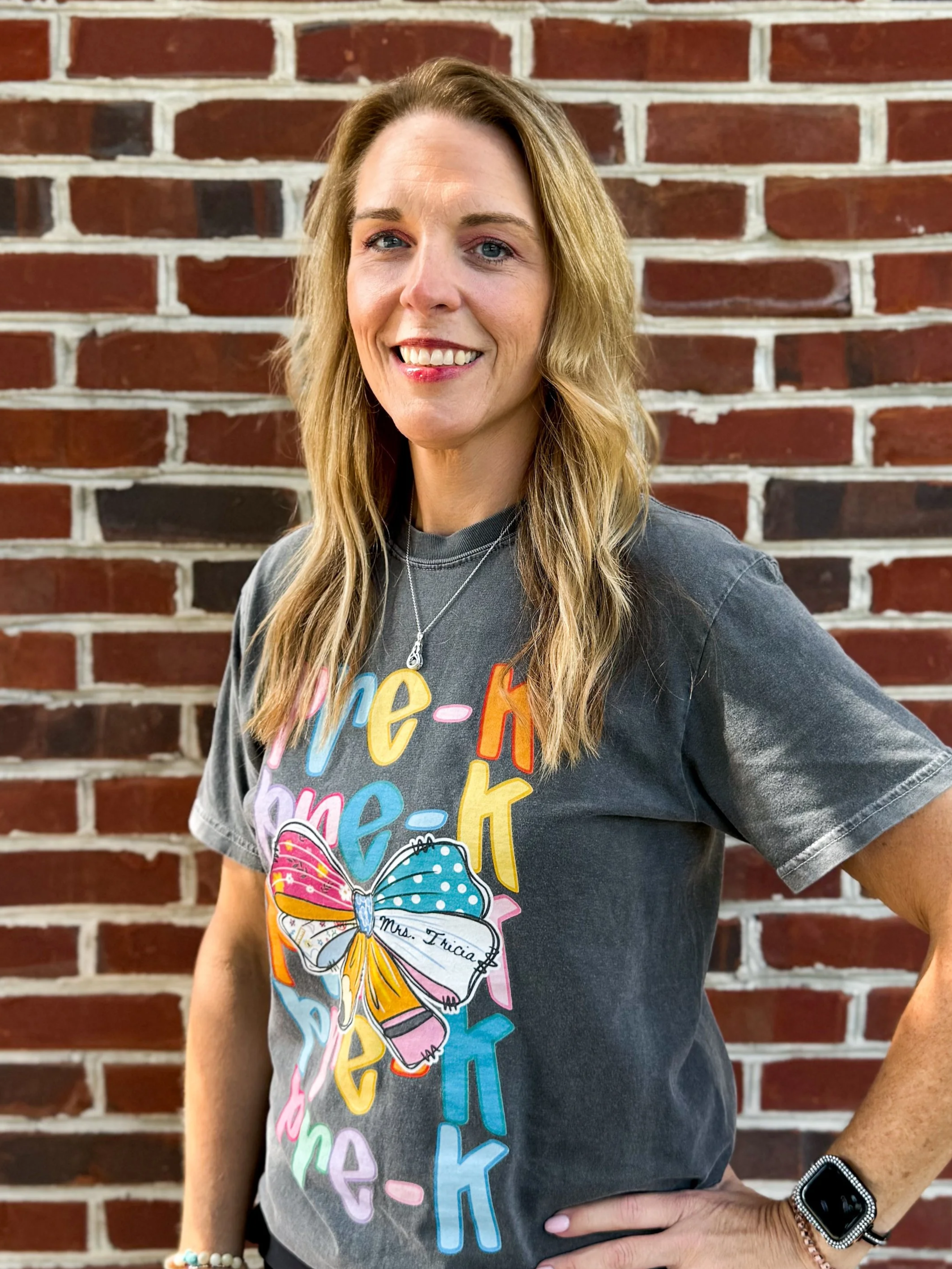 A smiling woman with blonde hair, wearing a dark gray t-shirt with colorful text and graphic design, standing in front of a brick wall.