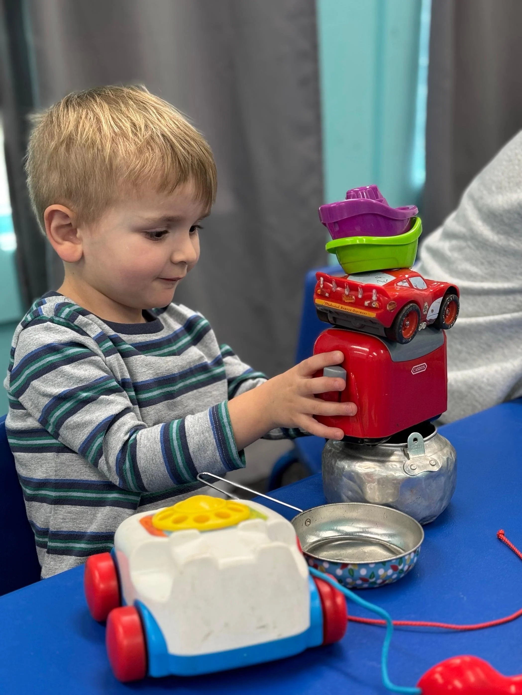 A young boy stacked toy cars and boats on top of a red toy cash register, with more toy items on a blue table, in a room with turquoise and gray walls.