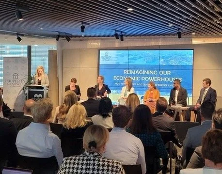 Panel discussion at a conference with a speaker at a podium and five panelists seated on stage, in front of an audience, with a large digital screen displaying 'Reimagining Our Economic Powerhouses' in the background.
