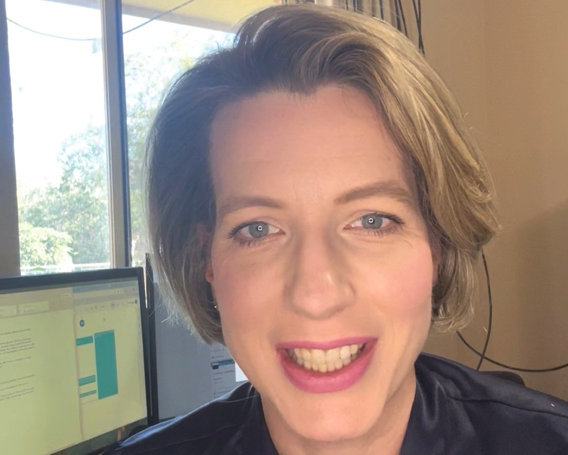 Close-up of a woman with short brown hair smiling at the camera, in an office with computers and a window showing trees outside.