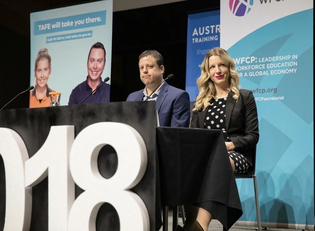 Three people are sitting at a panel discussion on stage during an event, with banners in the background related to workforce education and training.