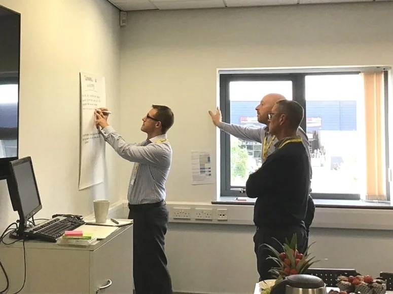 Three men in an office, one writing on a large sheet of paper on the wall, two observing, near a window with a view outside.