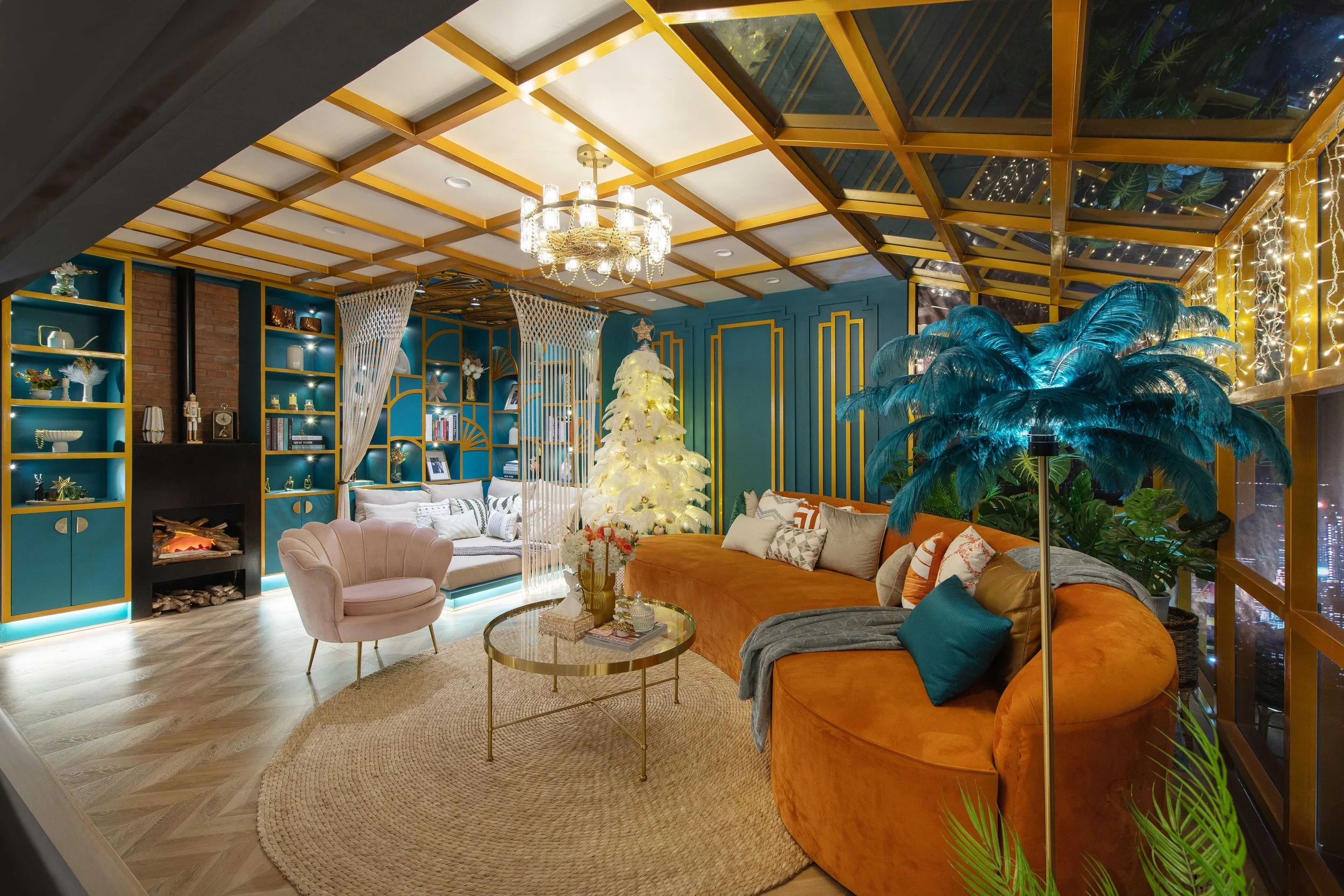 Cozy living room decorated for Christmas with a white Christmas tree, plush orange and gray sofas, blue accent wall with gold trim, bookshelves, and large indoor plants. A chandelier hangs from the ceiling and twinkling string lights are visible thro