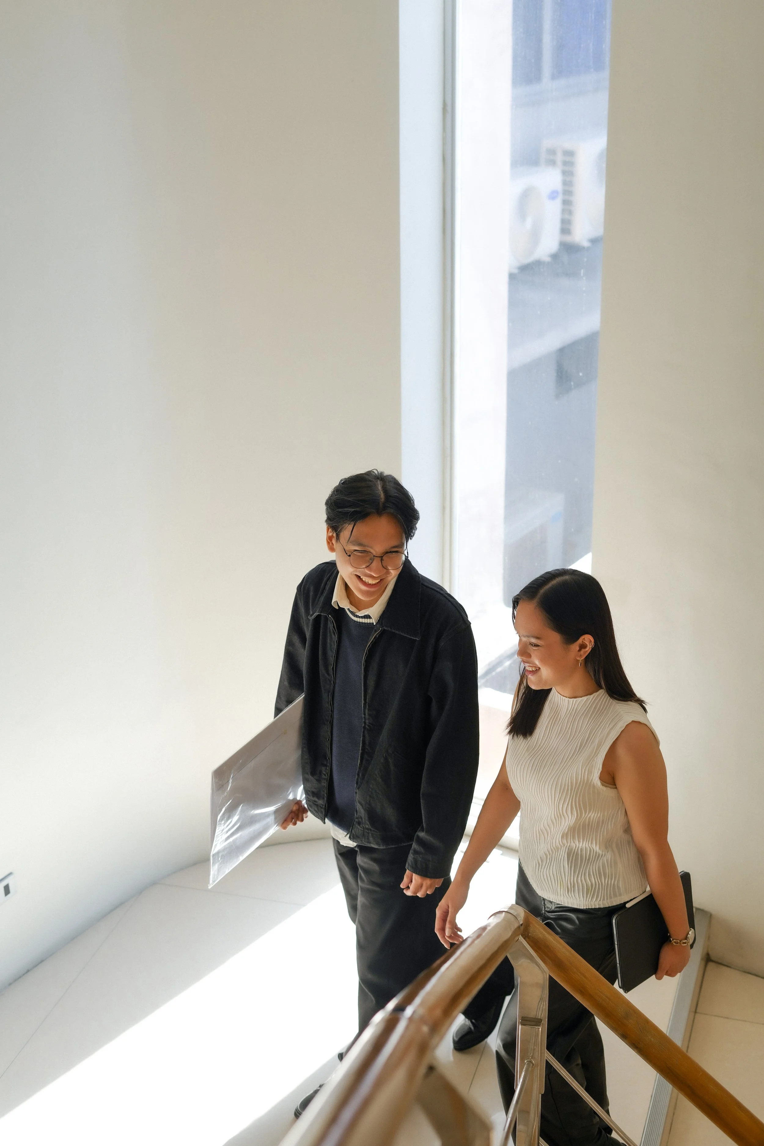 Two young adults, a man and a woman, are walking down a staircase by a large window. They are smiling, and the man is holding a large clear plastic bag. The woman is carrying a black folder. The scene is bright with natural light coming through the window.