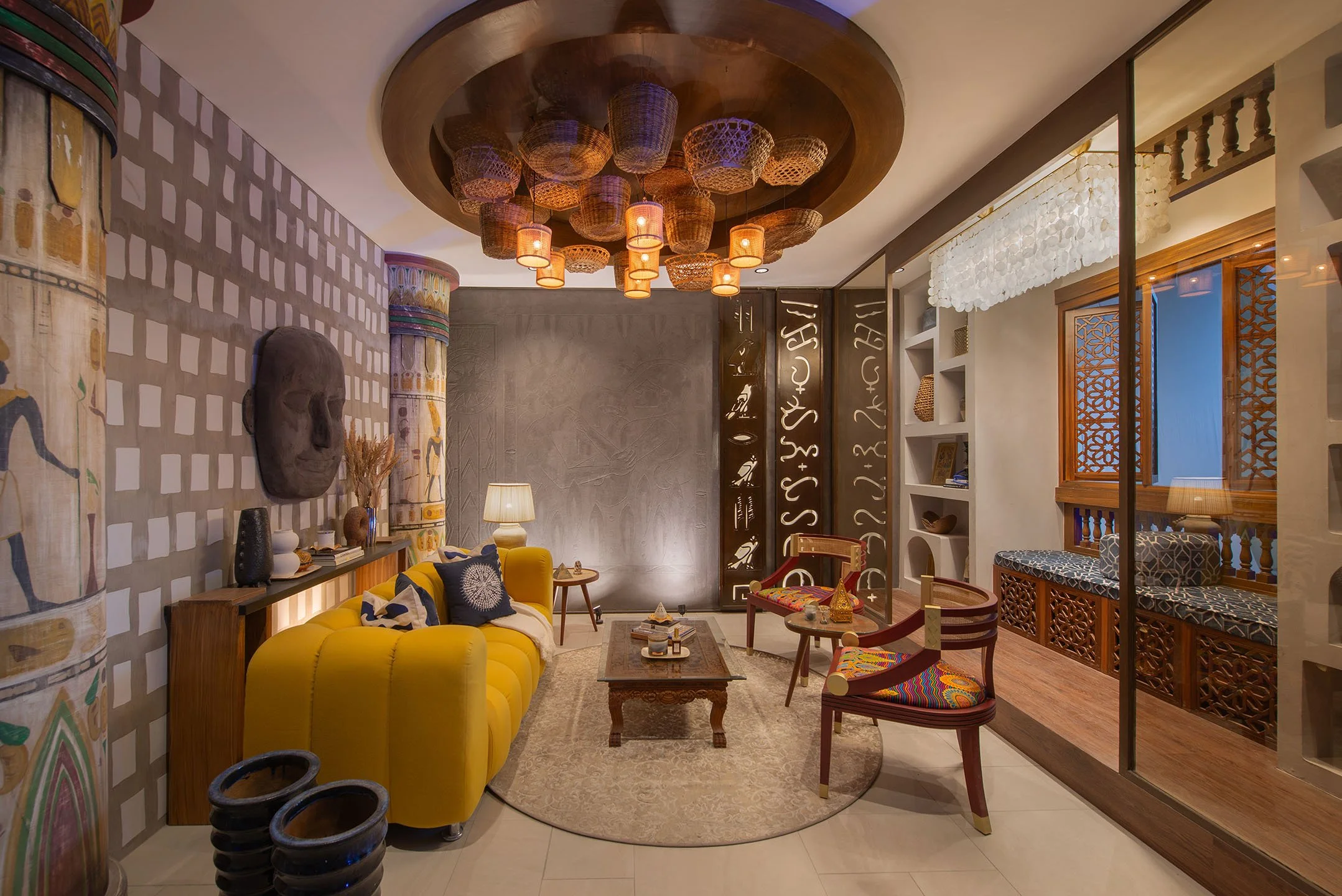 Interior of a living room with a yellow sofa, patterned cushions, wooden furniture, and decorative art, featuring hanging woven baskets from a wooden ceiling and wall art with Egyptian motifs and abstract lettering.