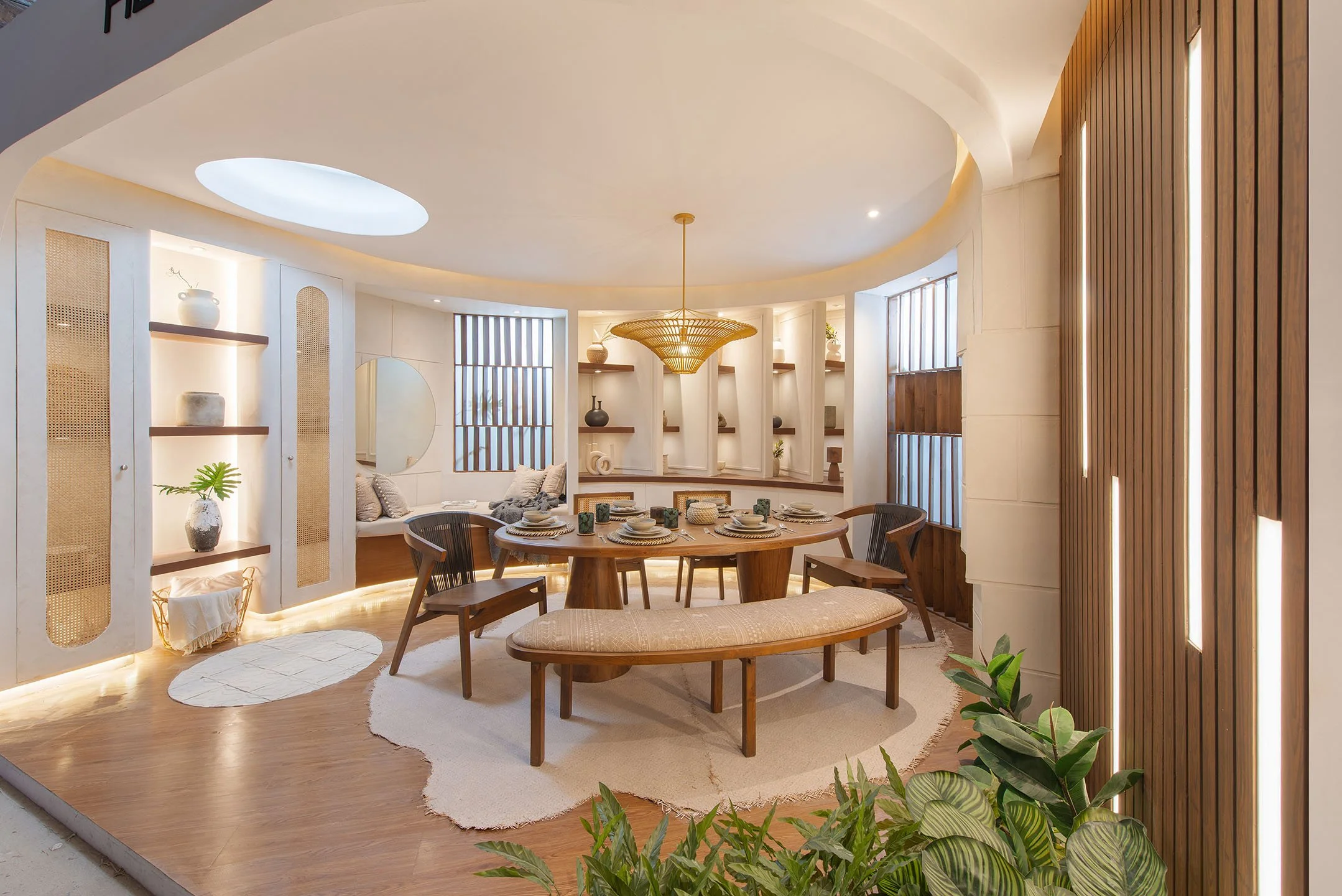A modern, well-lit living and dining room area with a round dining table set with plates, silverware, and cups, surrounded by wooden chairs. The room features built-in shelves with decorative items, a large circular mirror, a unique wooden pendant light, and warm lighting accents. There are large windows with striped blinds, a sconce, and indoor plants, creating a cozy and stylish ambiance.